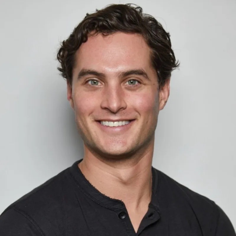 Smiling man with curly brown hair in a dark henley shirt, set against a plain light gray background, conveying a friendly and approachable demeanor.