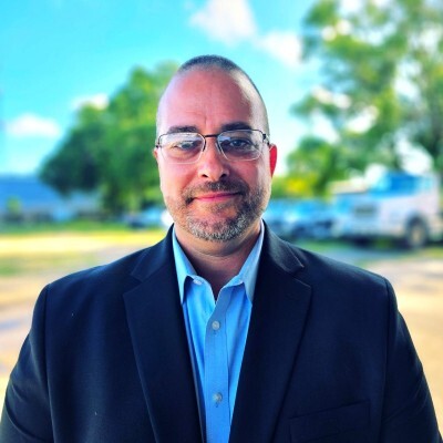 A man wearing glasses and a suit stands outdoors, smiling gently. The background features blurred greenery and a blue sky, conveying a calm, positive tone.
