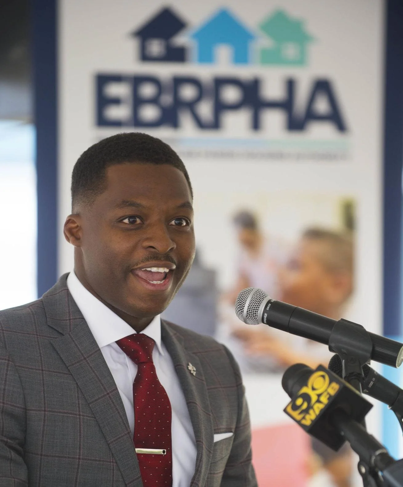 A man in a gray suit and red tie speaks energetically at a podium with a microphone. Behind him is a banner reading "EBRPHA."