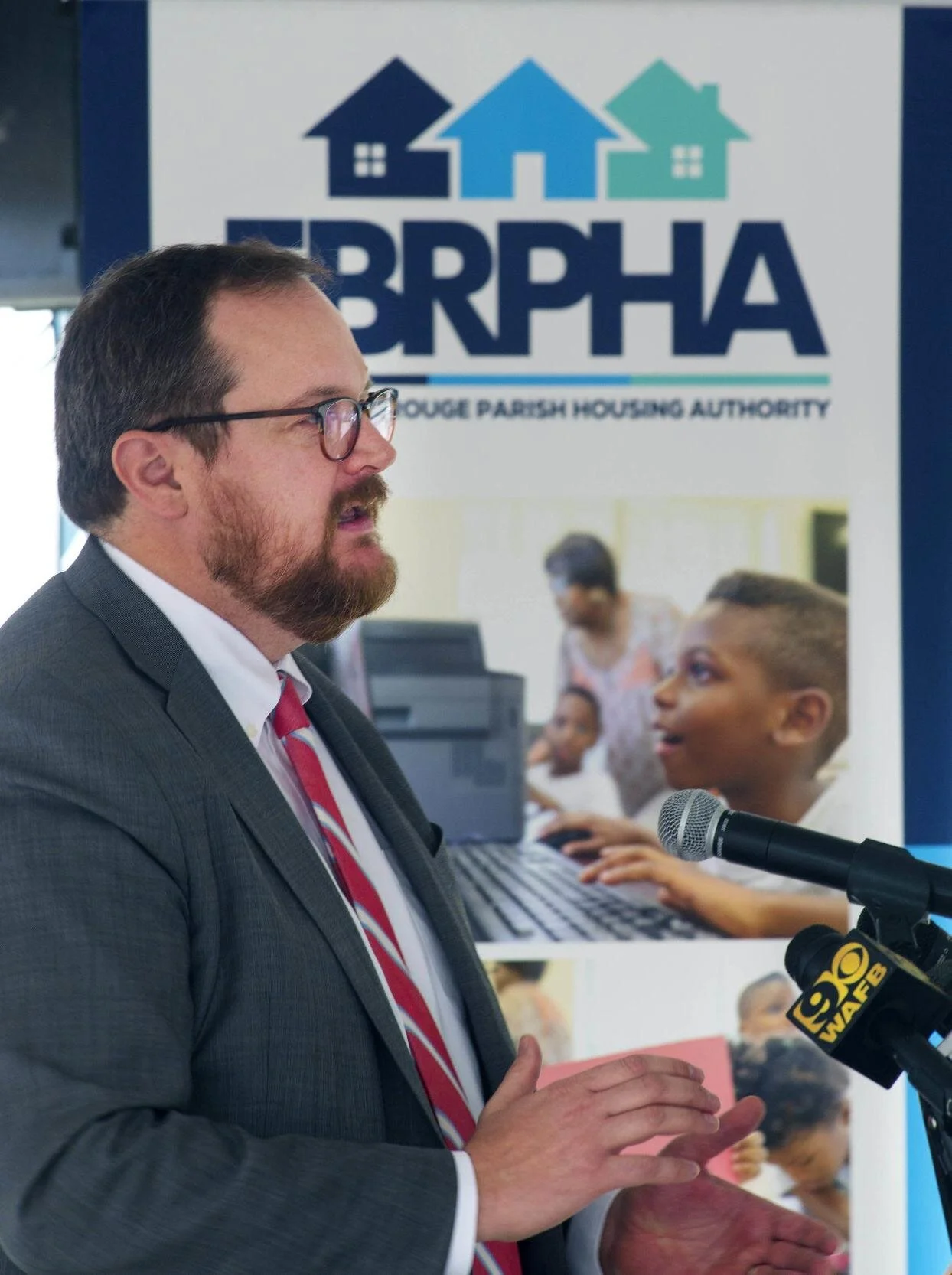 A man in a suit and red tie speaks at a podium with microphones. Behind him is a sign for the Baton Rouge Parish Housing Authority, featuring a child using a computer.