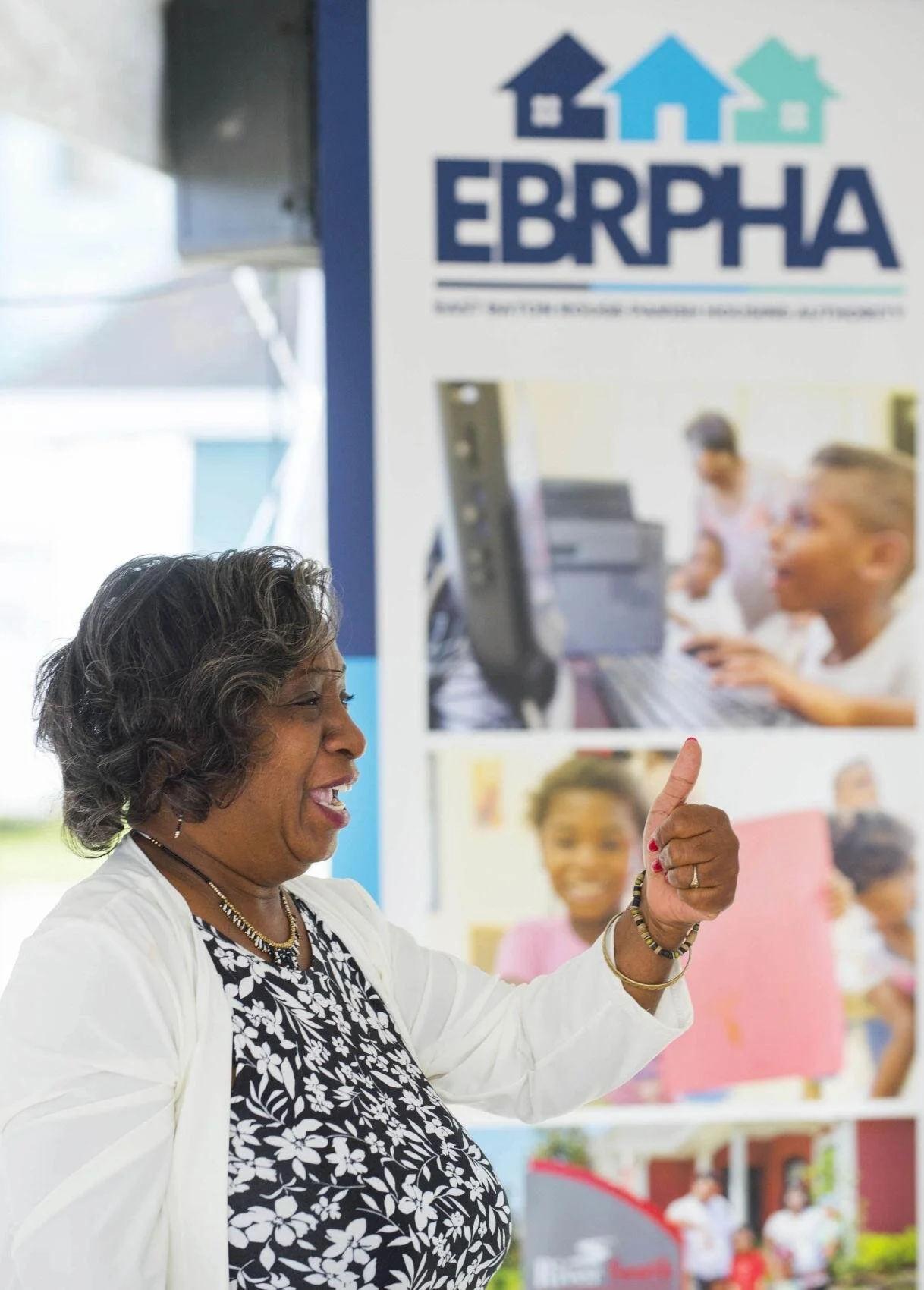 A woman smiles and gestures a thumbs-up in front of an EBRPHA banner with photos of children using computers, conveying a positive and supportive tone.