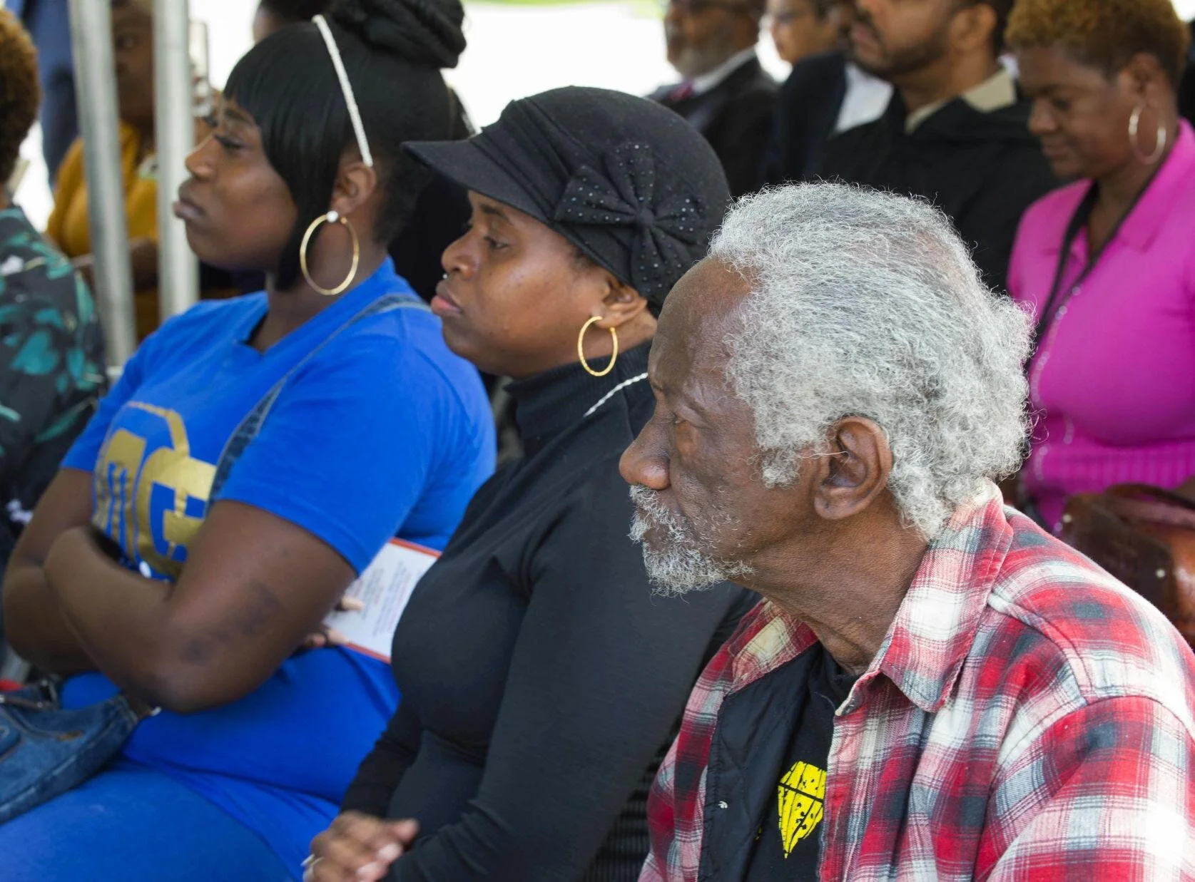 A diverse group of people sit attentively at an outdoor event. The foreground focuses on an elderly man with gray hair in a plaid shirt, conveying a contemplative mood.