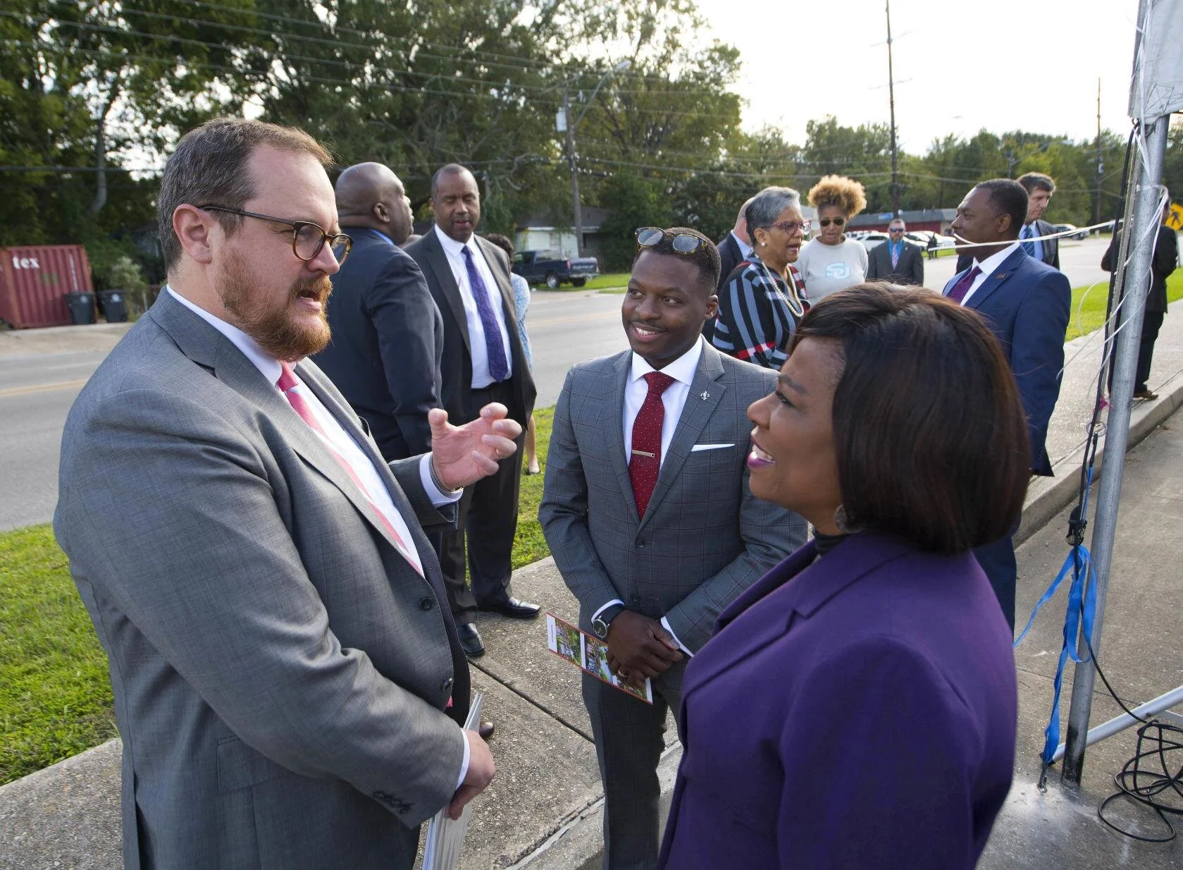 A group of people in formal attire engage in a lively outdoor conversation. The scene is positive, with smiling faces, set on a sunny day.