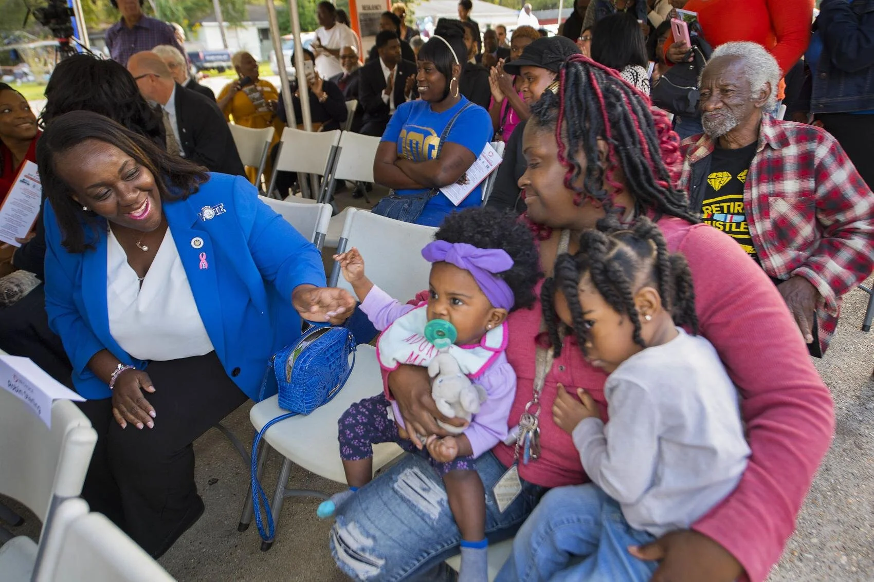 A joyful woman in a blue blazer interacts with a mother holding two young children at an outdoor gathering. Onlookers sit on chairs, creating a warm, community-focused atmosphere.