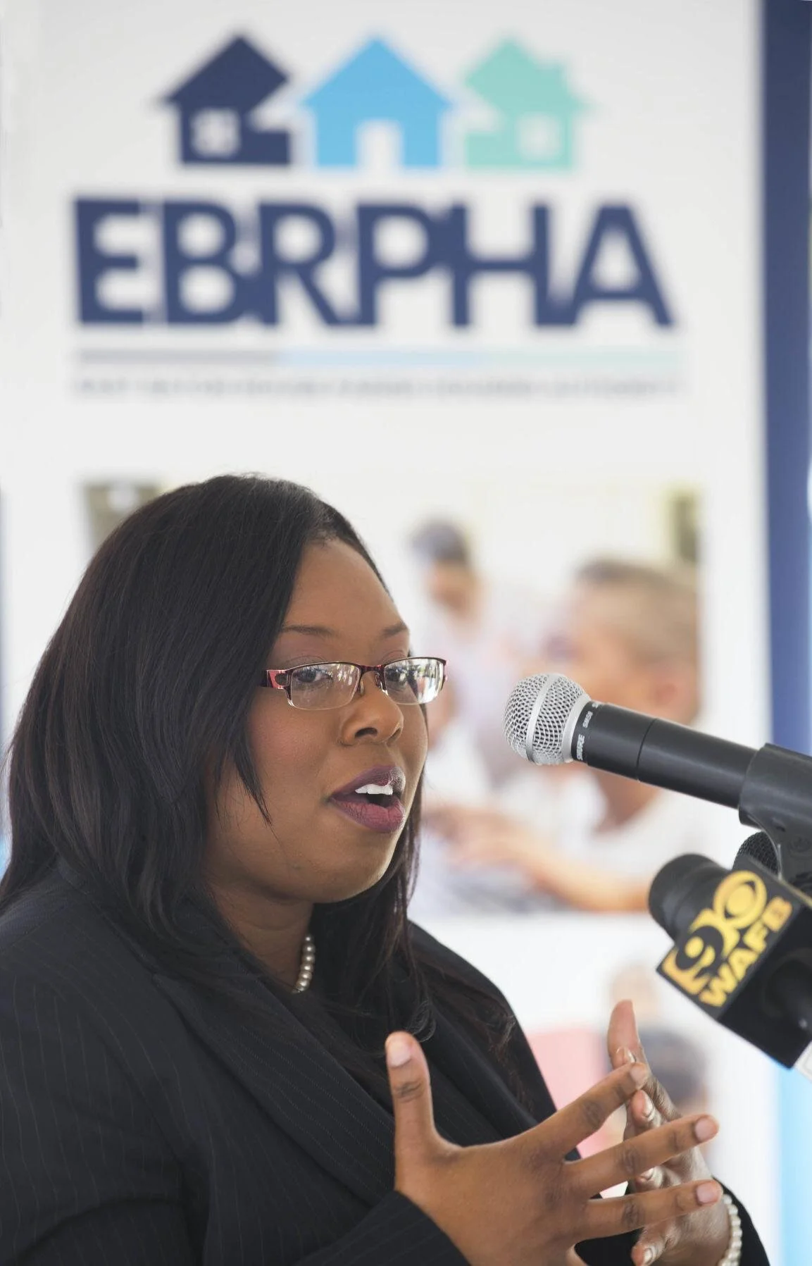A woman in glasses speaks at a podium with a microphone, gesturing confidently. Behind her is an EBRPHA banner with house icons, suggesting a formal event.
