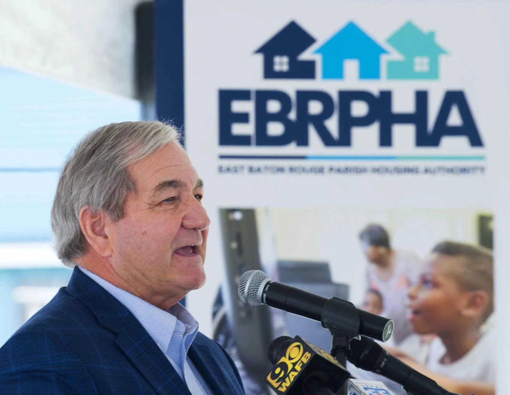 A person speaks at a podium with a microphone. Behind them is the EBRPHA logo and a blurred image of children. The setting is professional and engaging.