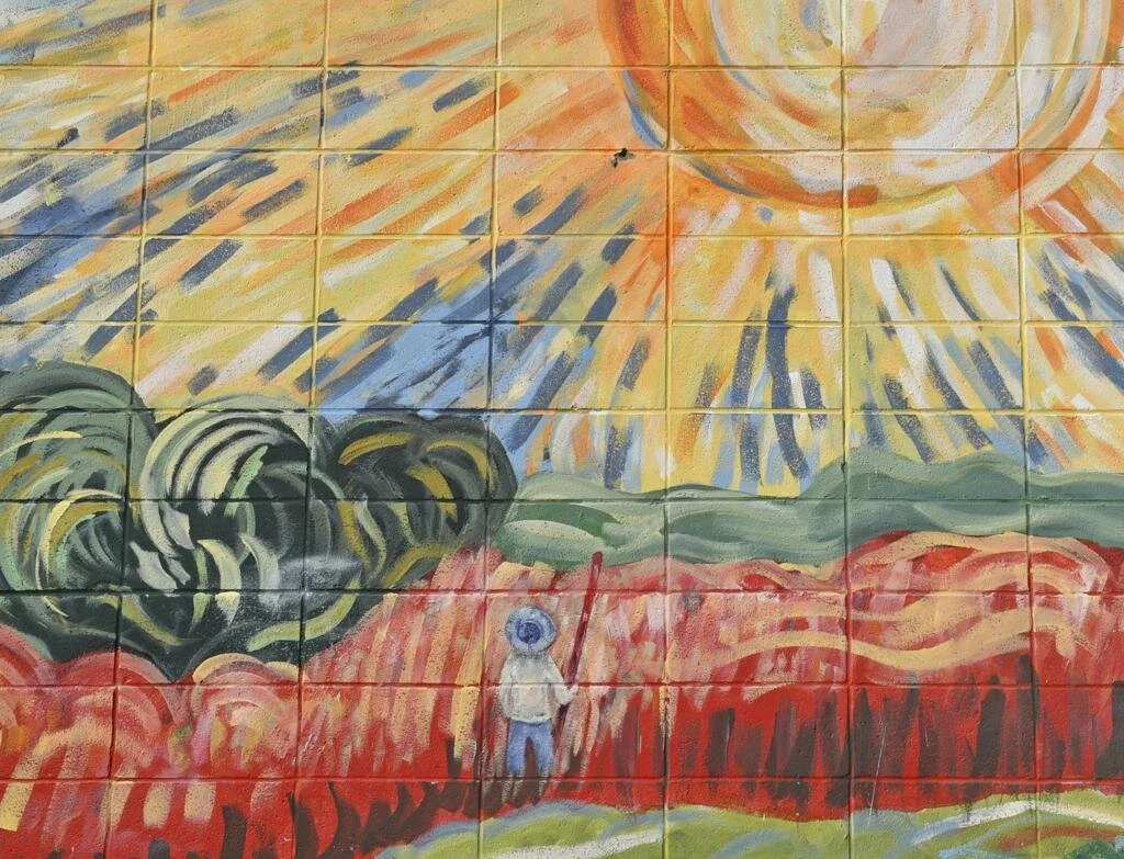 Mural of a bright sun radiating over a field with a solitary figure in a hat holding a stick. Vibrant colors create a warm, hopeful tone.