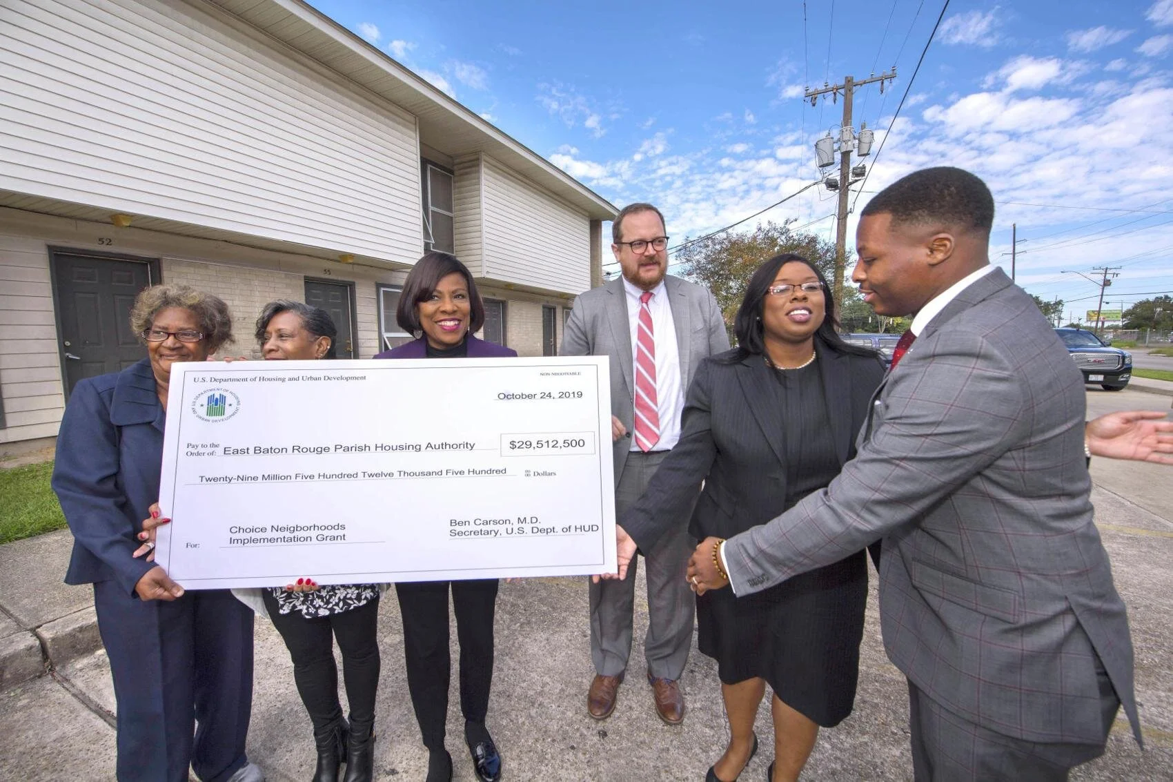 A diverse group of people stands outdoors holding a large check for $29,512,500, smiling and appearing celebratory. The background shows a residential building and a clear sky.