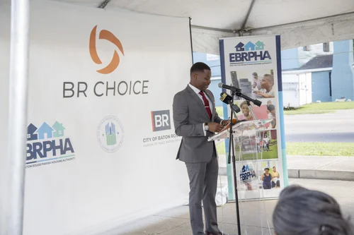 Man in suit speaks at podium with BR Choice and Housing Authority logos in background.
