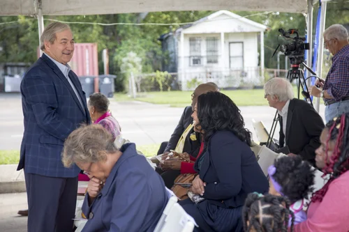 Man in a blue suit jacket smiles while greeting seated attendees under a tent.