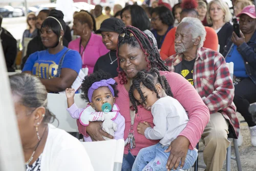 Woman holds two toddlers while seated among a diverse crowd at a community event.