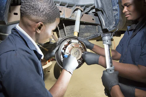 Two students work on a vehicle's rear drum brake assembly, with one using a tool for adjustment while the other holds a work light.