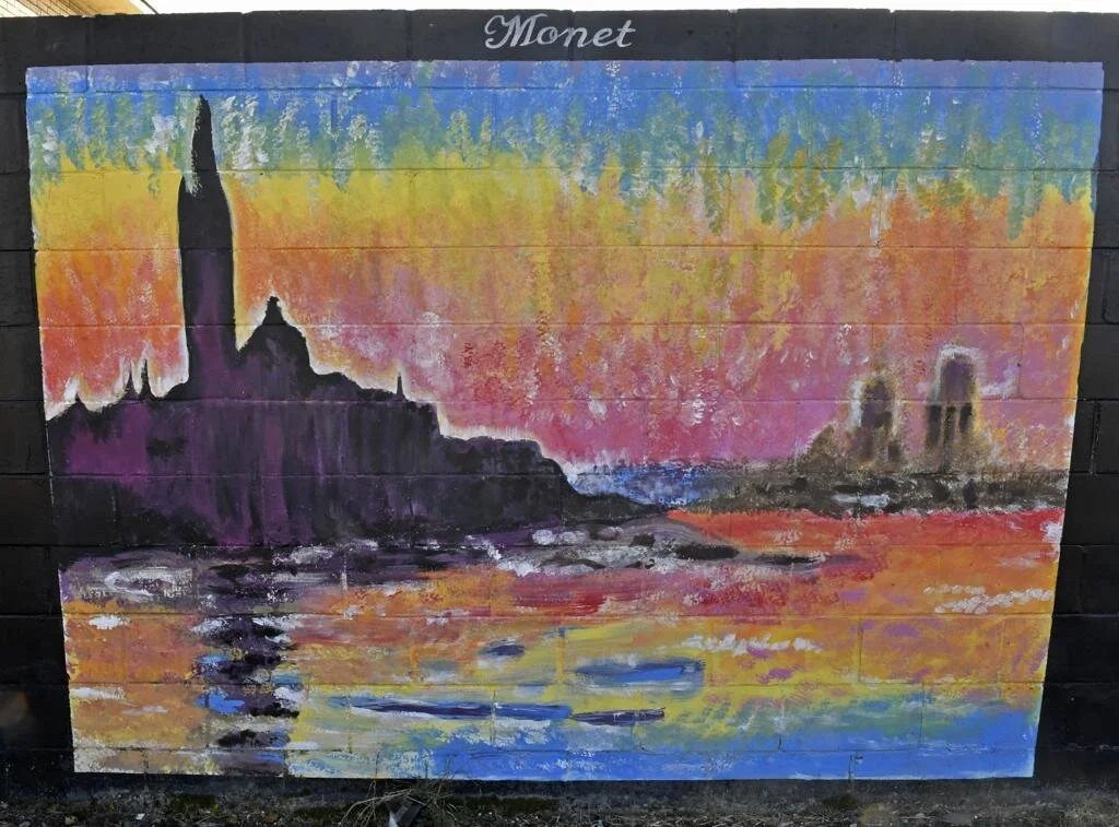 Mural painted on a brick wall depicting a colorful sunset over water with dark silhouettes of buildings, inspired by Monet's style. The word "Monet" is written at the top.