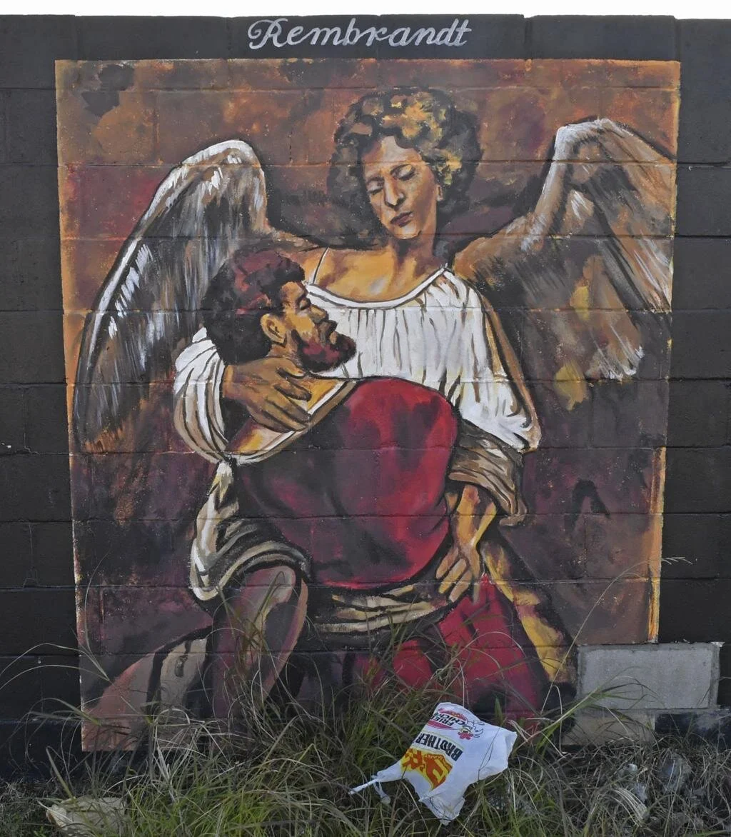 A mural on a brick wall depicts an angel embracing a man, both draped in robes. The scene conveys warmth and comfort. "Rembrandt" is written above.