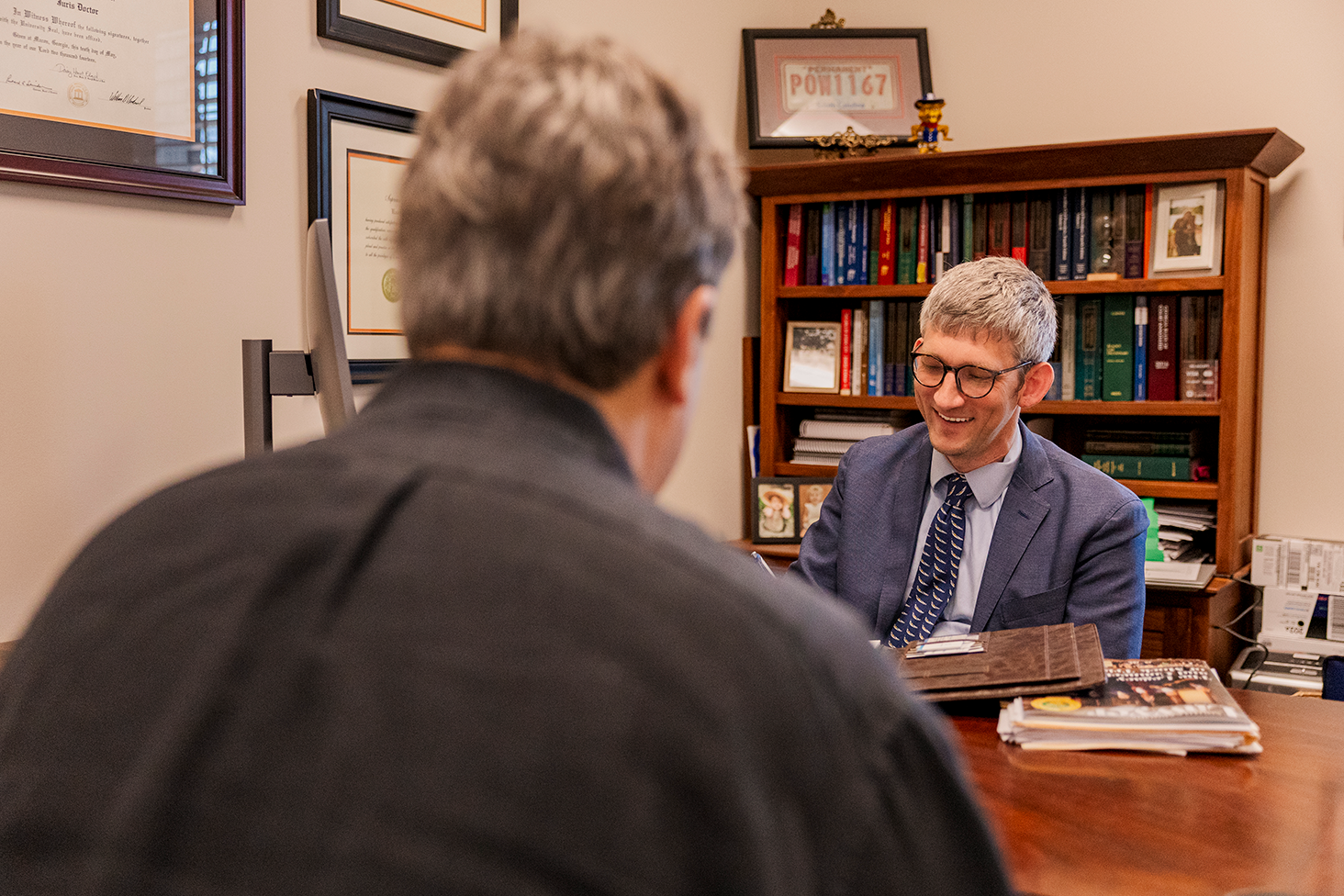 Smiling man in glasses and suit sitting at desk in an office, talking to another person.