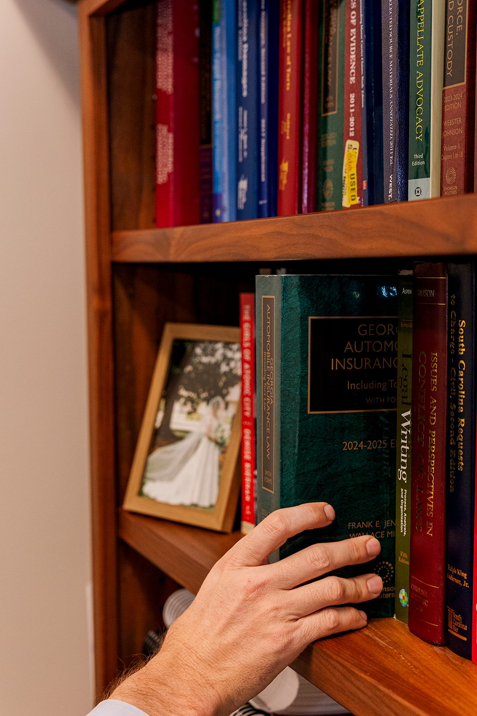 Hand pulling a green book titled 'Georgia Automobile Insurance Law' from a wooden bookshelf filled with legal books and a framed wedding photo.
