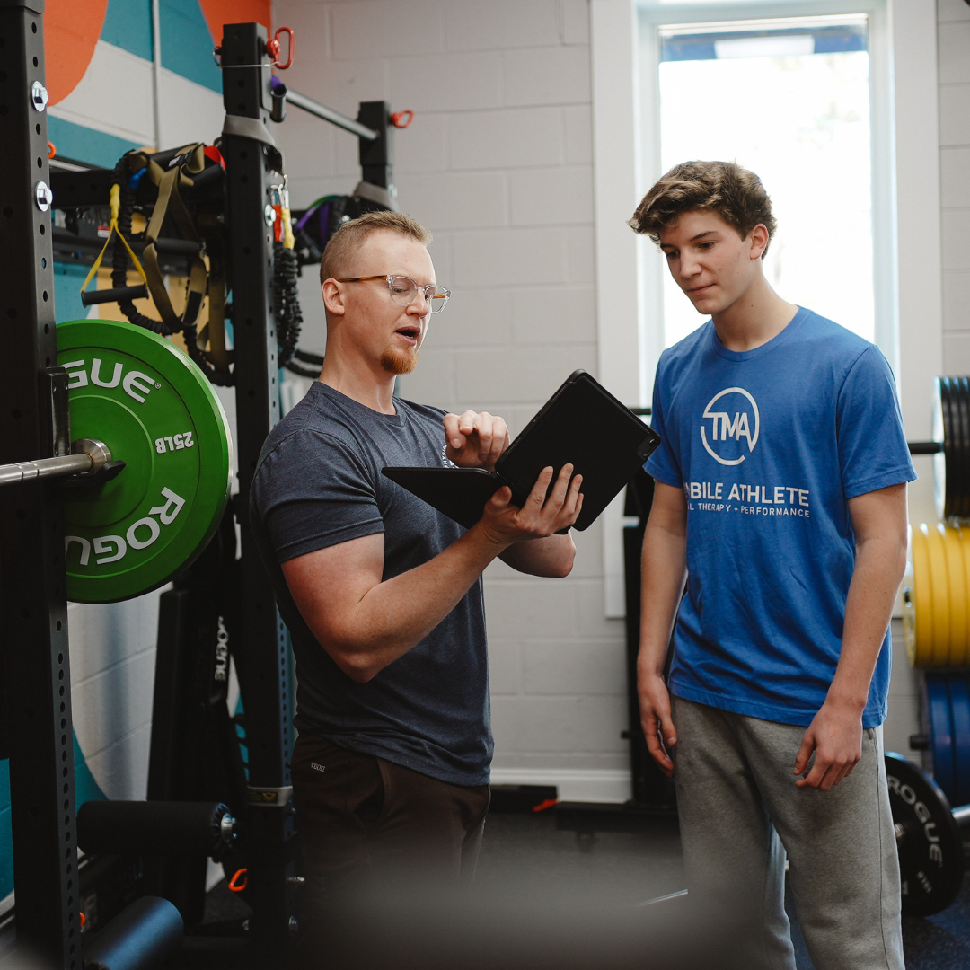 Fitness coach explaining workout details on a tablet to a young male athlete in a gym setting.