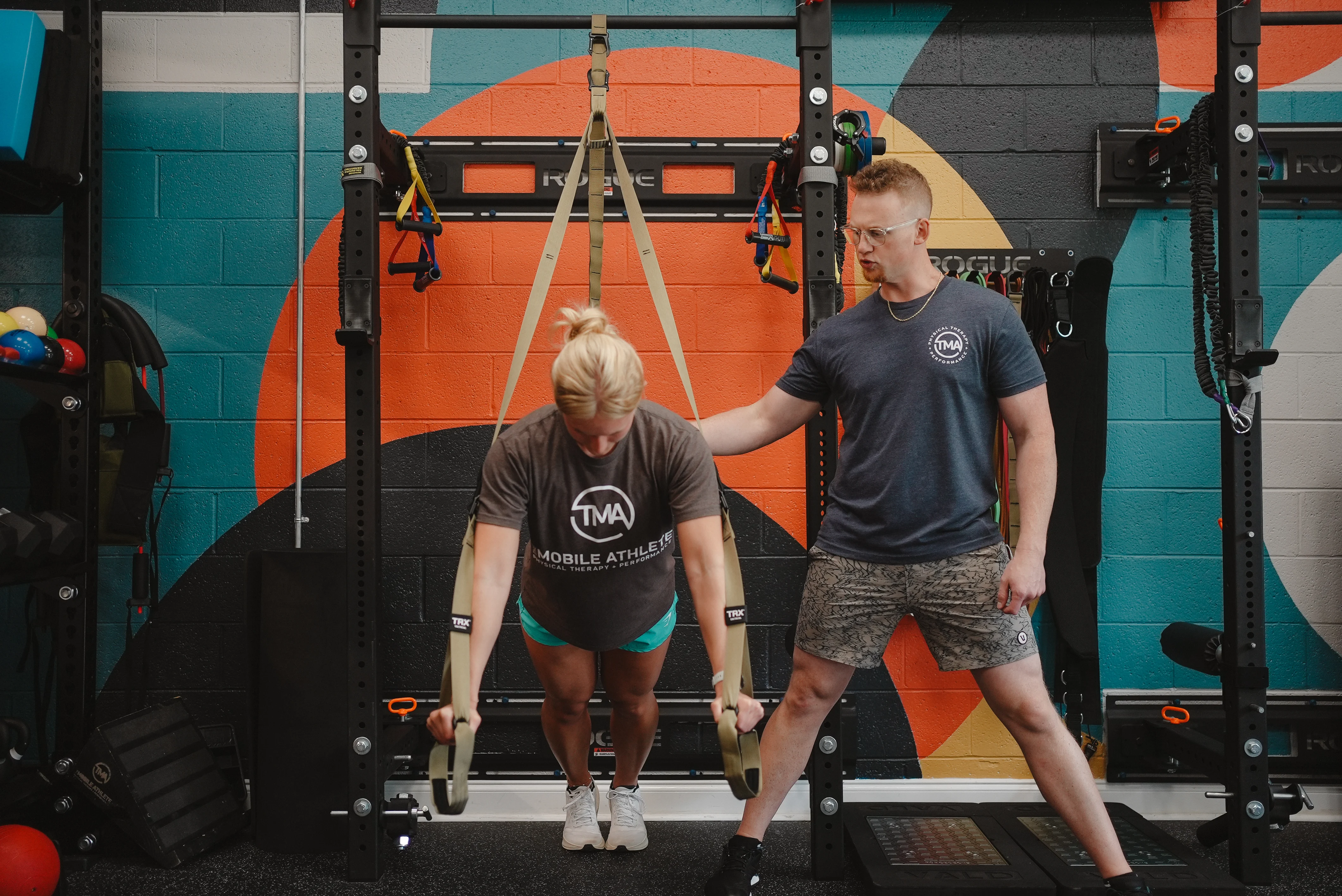 Trainer guides a woman performing a TRX suspension exercise in a gym with colorful geometric wall art.