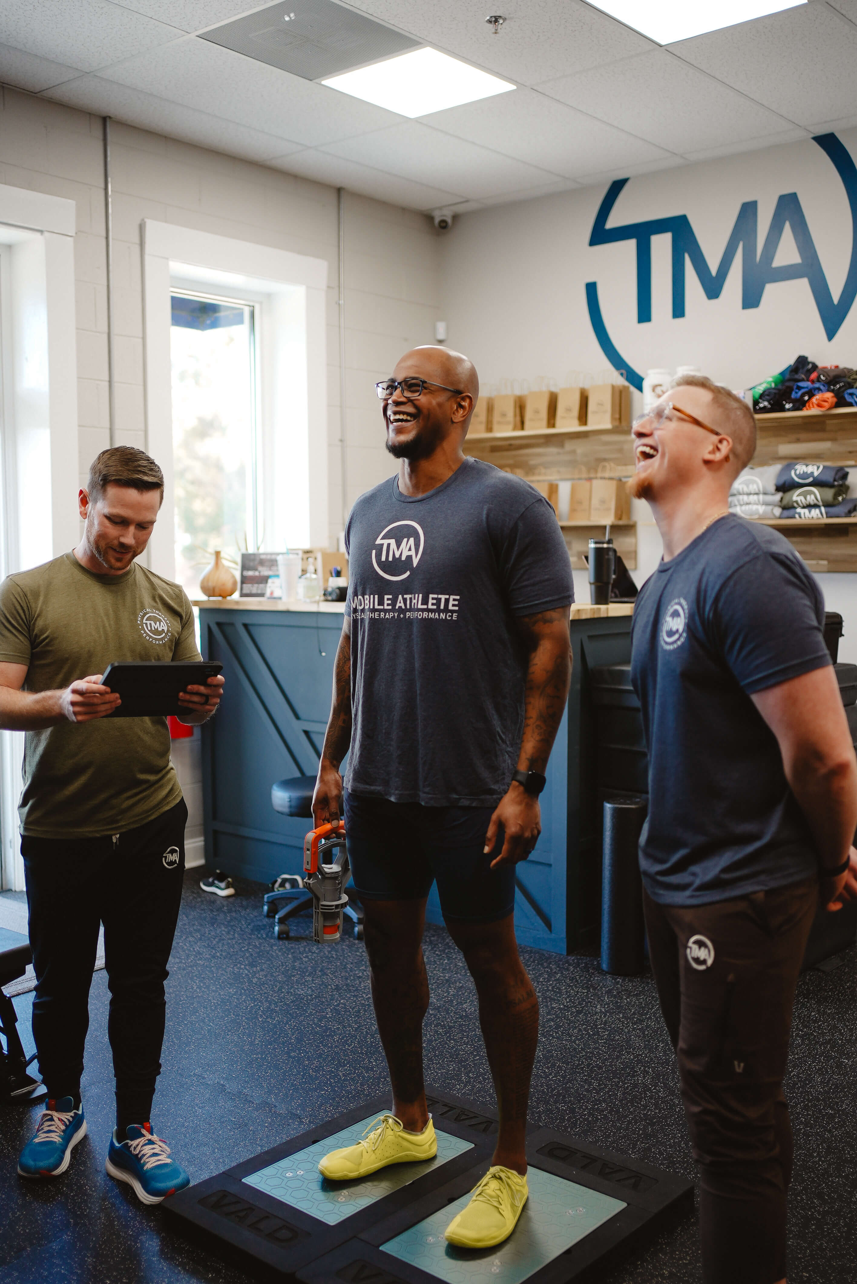 Three men in a fitness studio, two wearing TMA shirts, one holding a tablet, another standing on a VALD balance platform holding a device, all smiling and interacting.