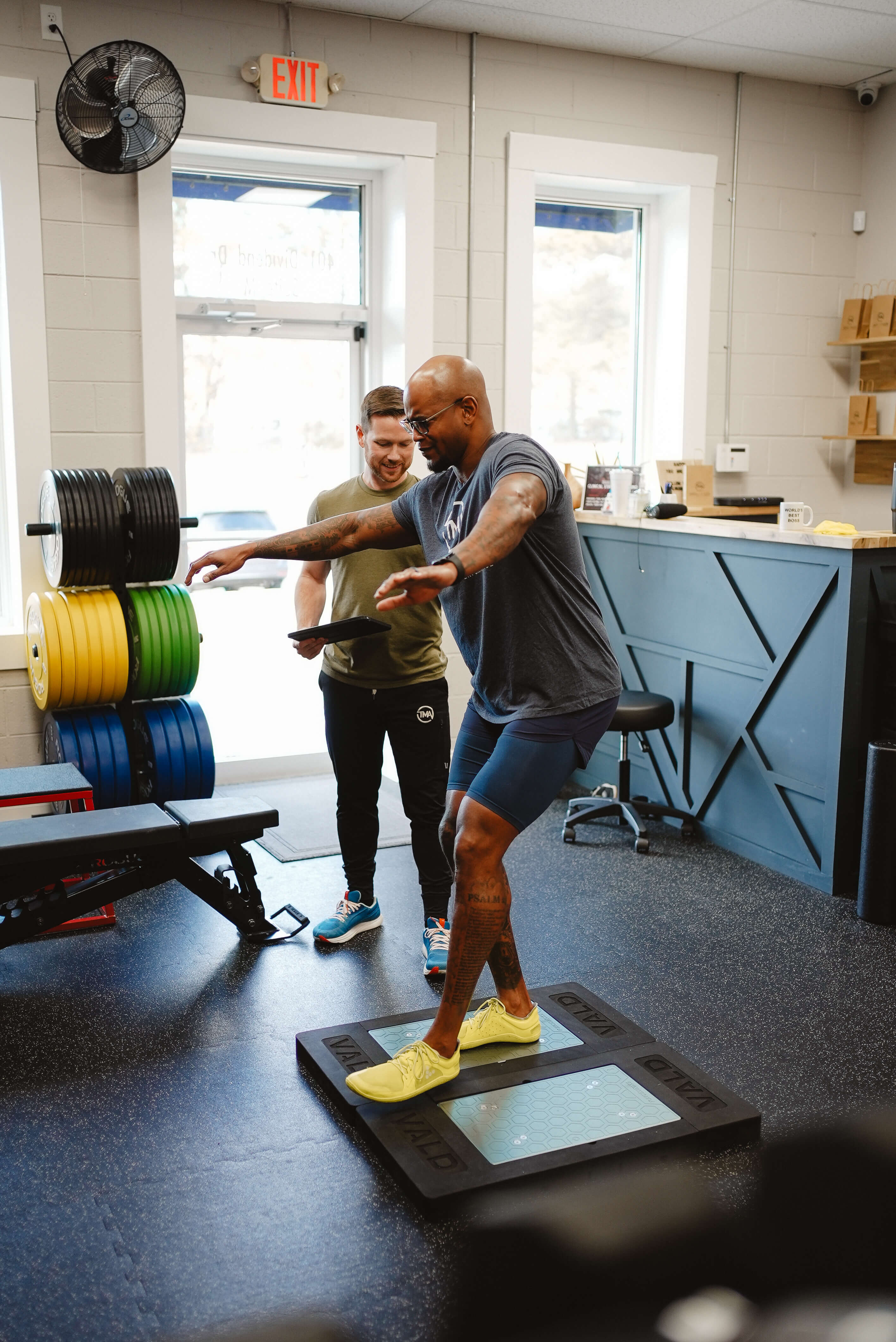 Man in athletic wear balancing on a balance board with arms extended while trainer watches and holds a tablet in a gym.
