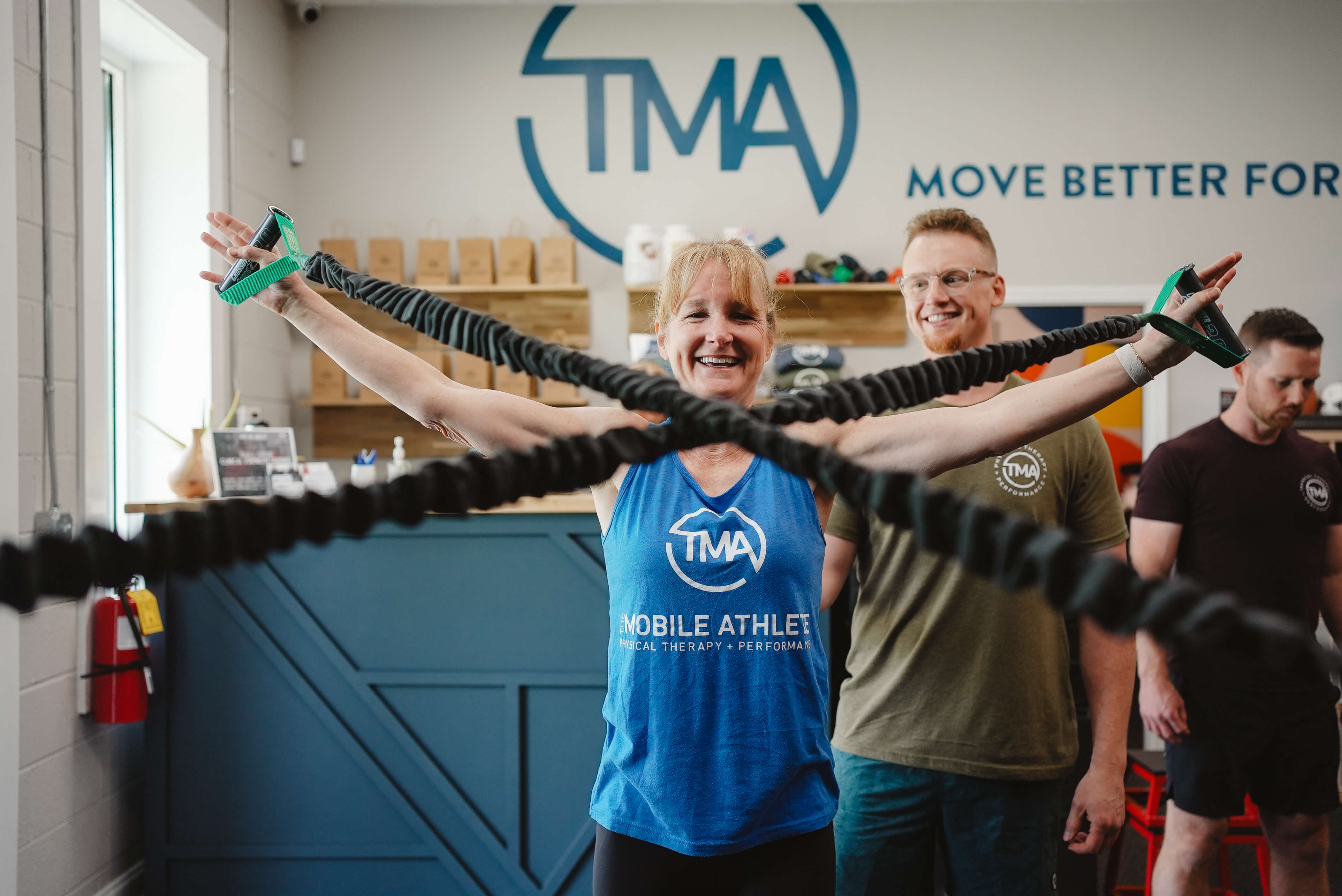 Smiling woman exercises with resistance bands stretched wide, assisted by two men in a physical therapy center.