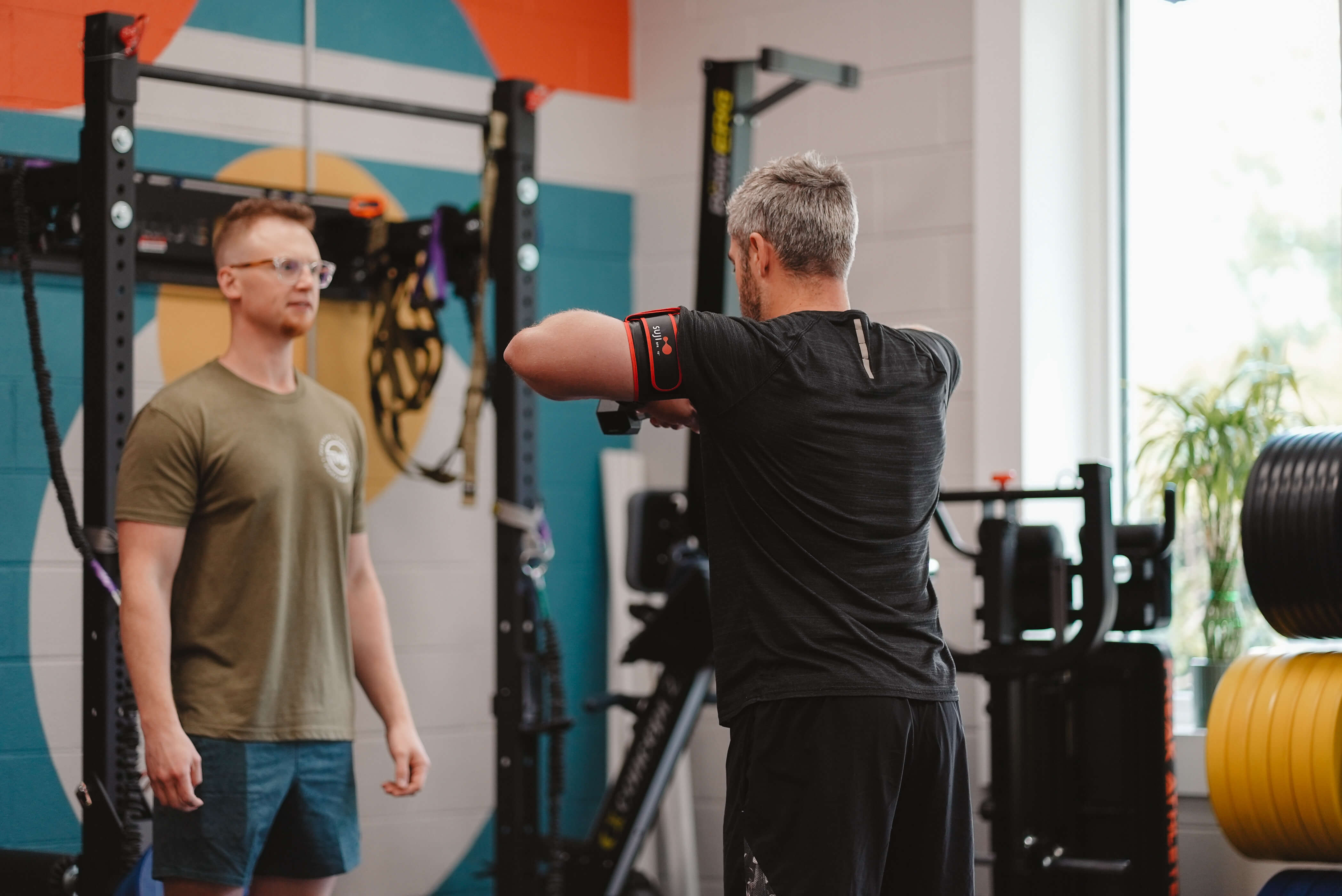 Two men in a gym; one wearing glasses in a green shirt watches another man exercising with arms raised and a fitness tracker on his upper arm.