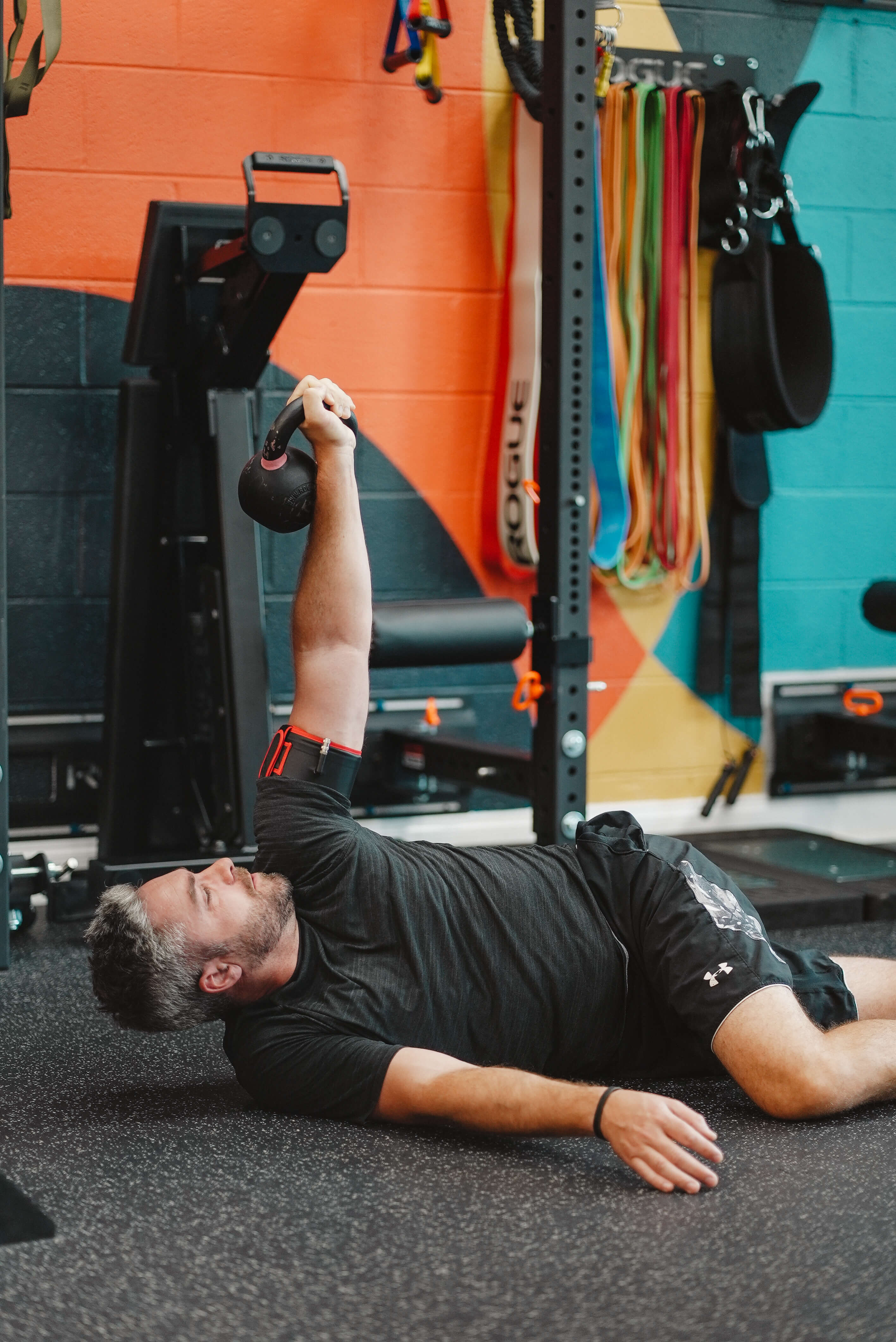 Man lying on gym floor lifting a kettlebell overhead with one arm during workout.