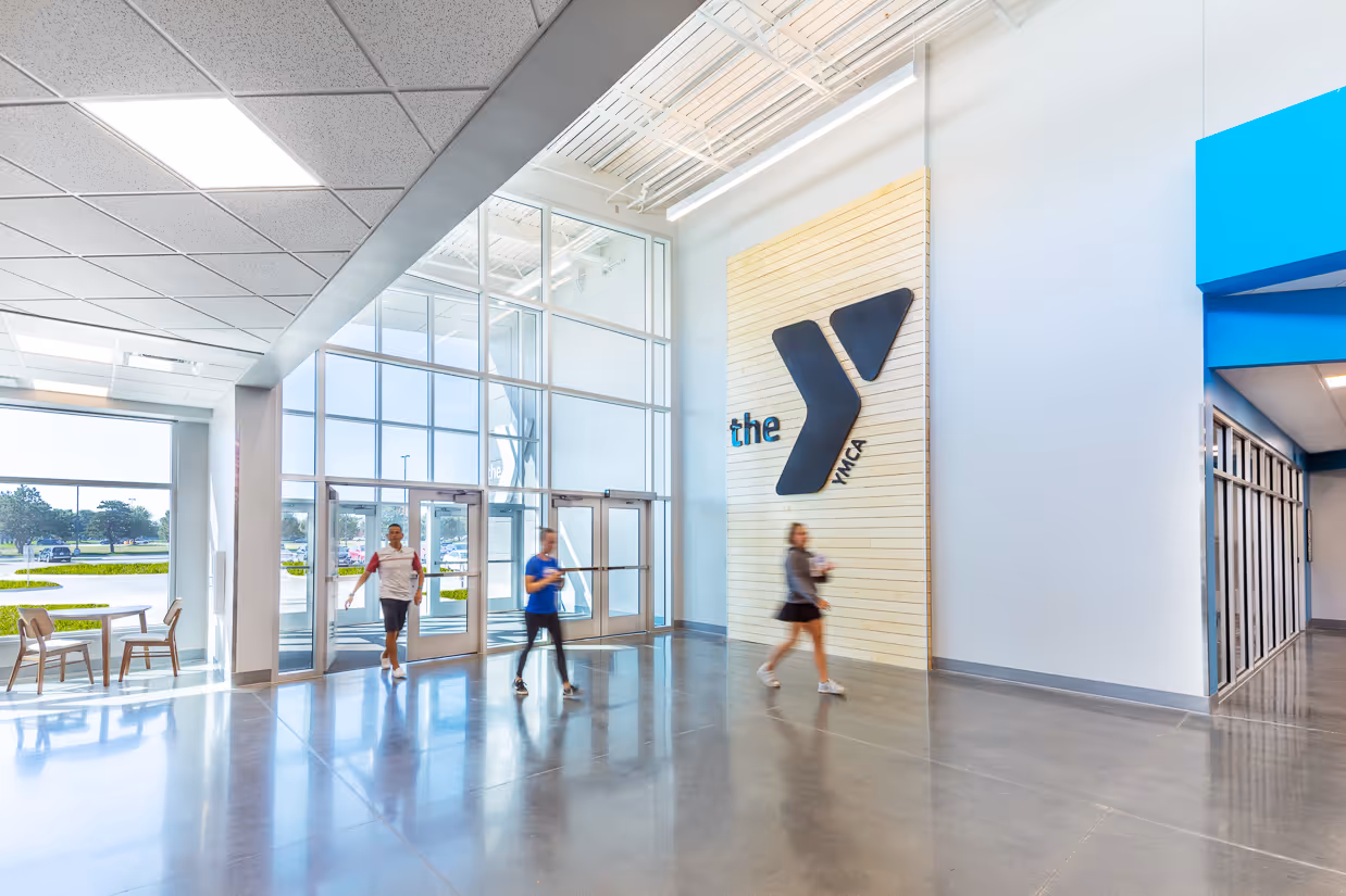 Bright YMCA lobby with large glass entrance doors, polished concrete floor, and three people walking inside.