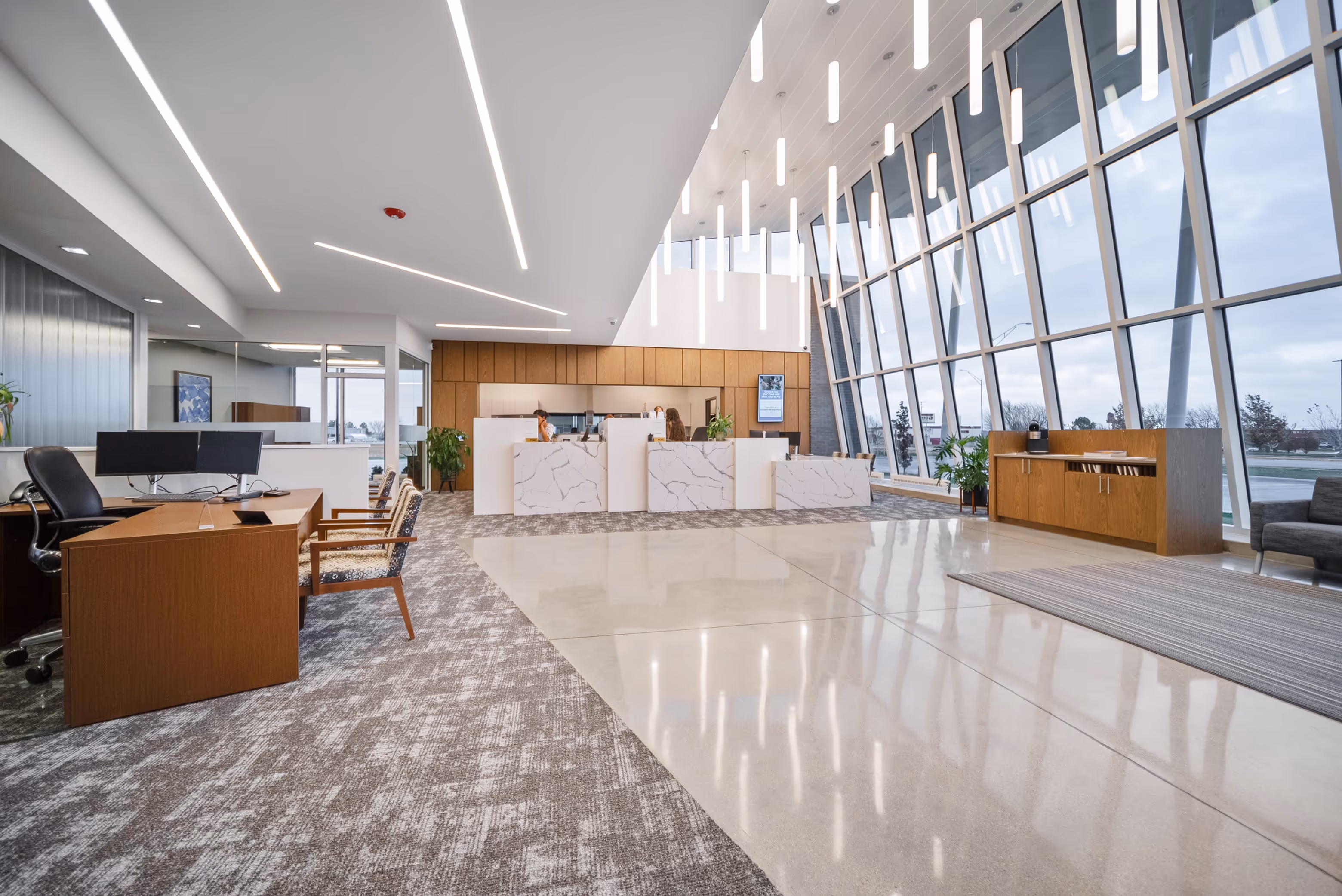 Modern office reception area with marble front desks, wood accents, large slanted glass windows, and hanging tube lights.