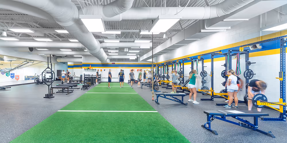 Indoor fitness gym with green turf in the center, weightlifting racks along the right wall, and several people exercising.