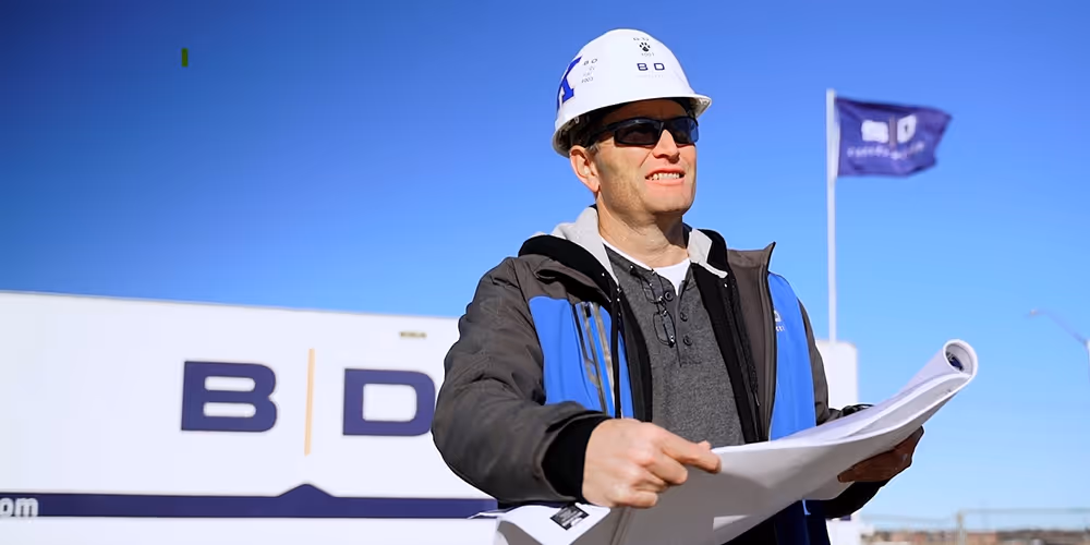 Construction worker wearing a hard hat and sunglasses holding blueprints outdoors with a B|D sign and flag in the background.