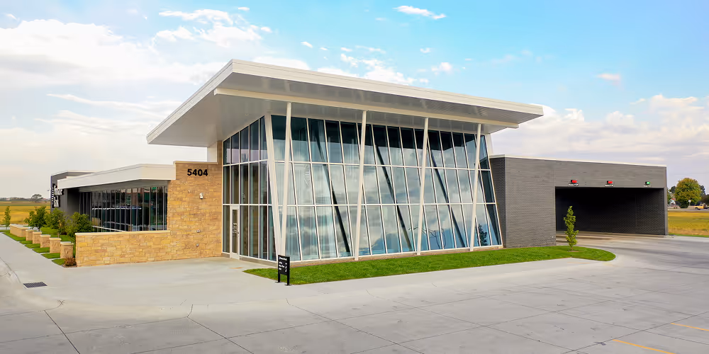 Modern commercial building with large angled glass windows, stone and dark gray walls, and a car drive-thru lane on the right.