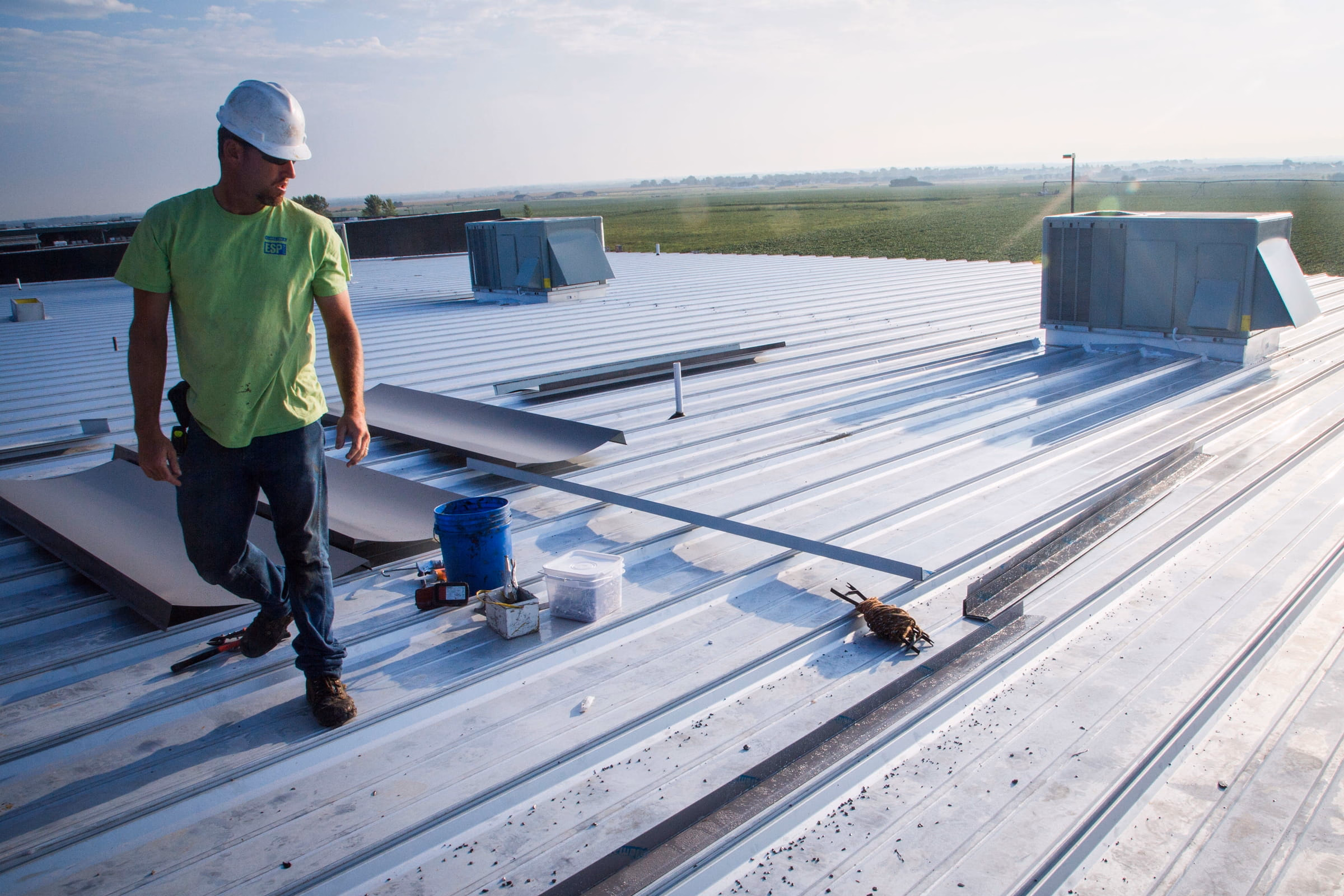 Construction worker in a green shirt and white hard hat standing on a metal rooftop with tools and equipment scattered nearby.