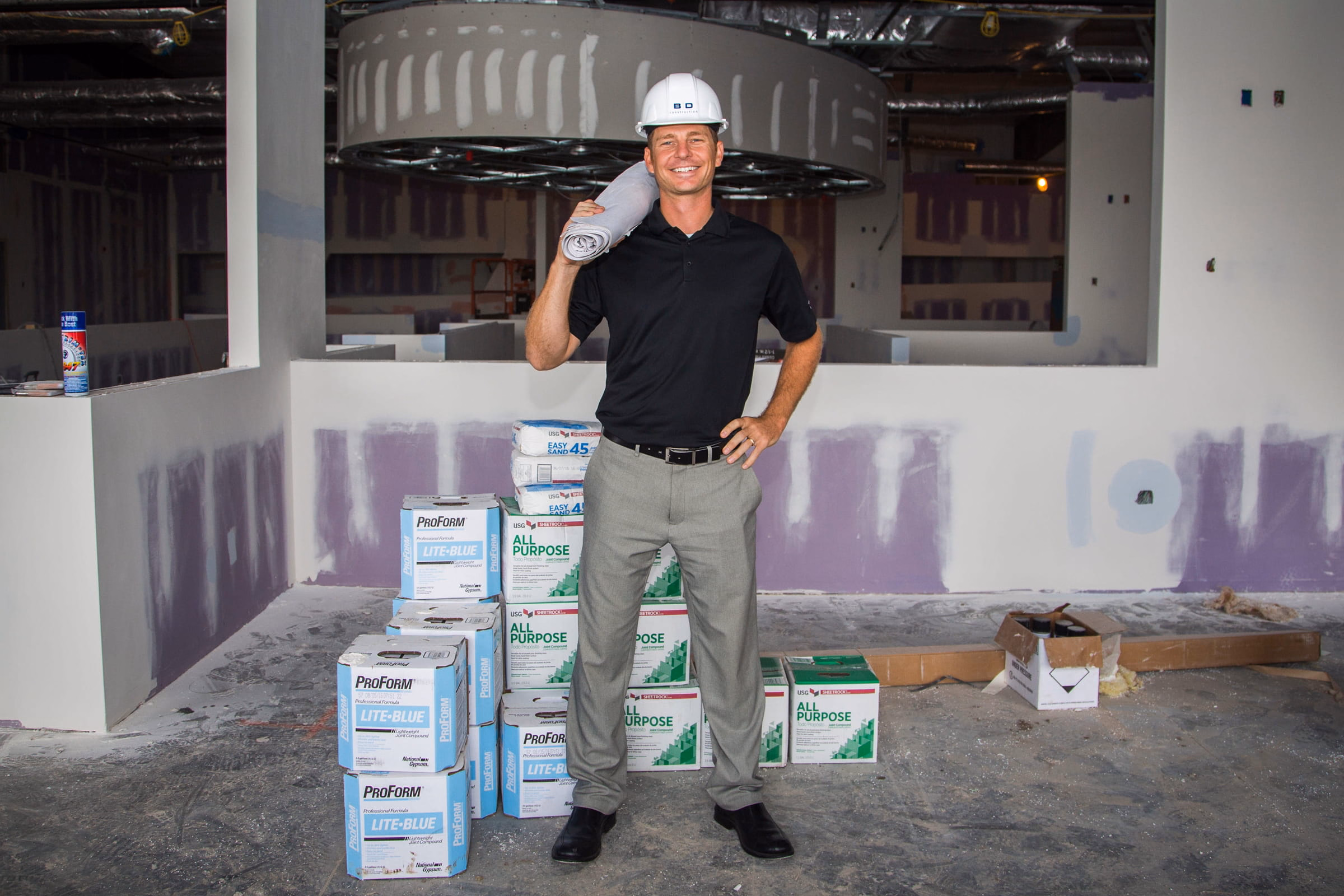 Smiling construction worker in a white hard hat and black shirt holding a rolled-up plan on his shoulder, standing inside an unfinished building with stacks of construction materials around.