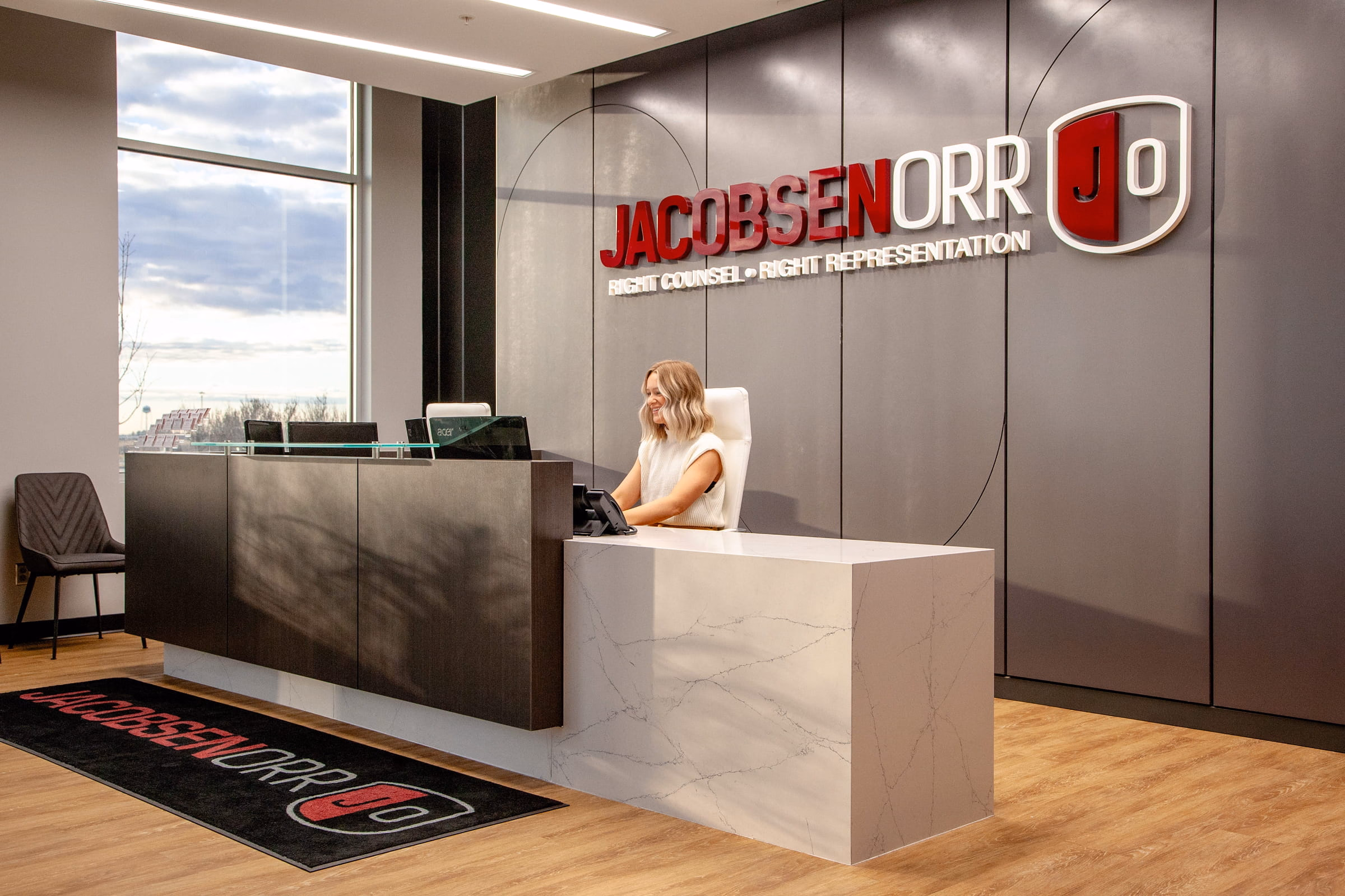 Reception desk with a smiling woman seated, with Jacobsen Orr logo and slogan on the wall behind her.