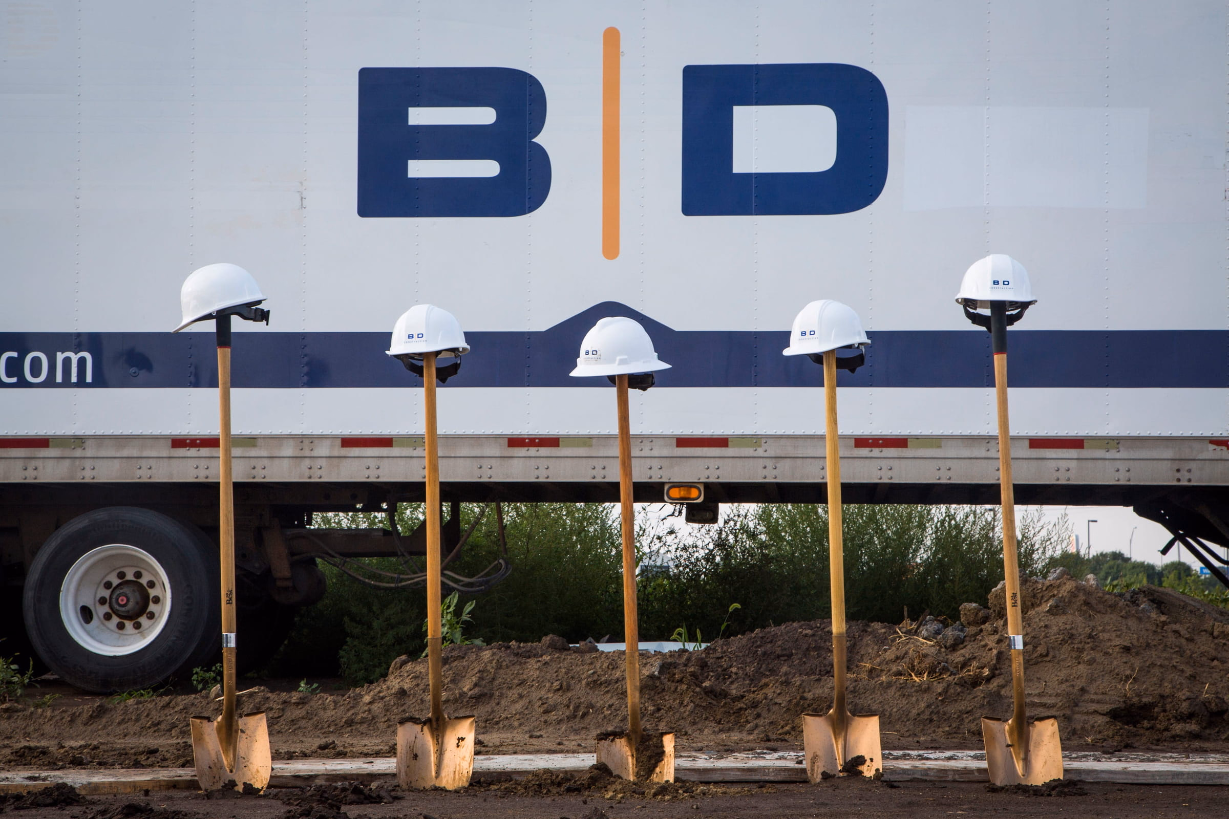 Five shovels with white hard hats on top, stuck in dirt in front of a large truck with the letters B and D on its side.