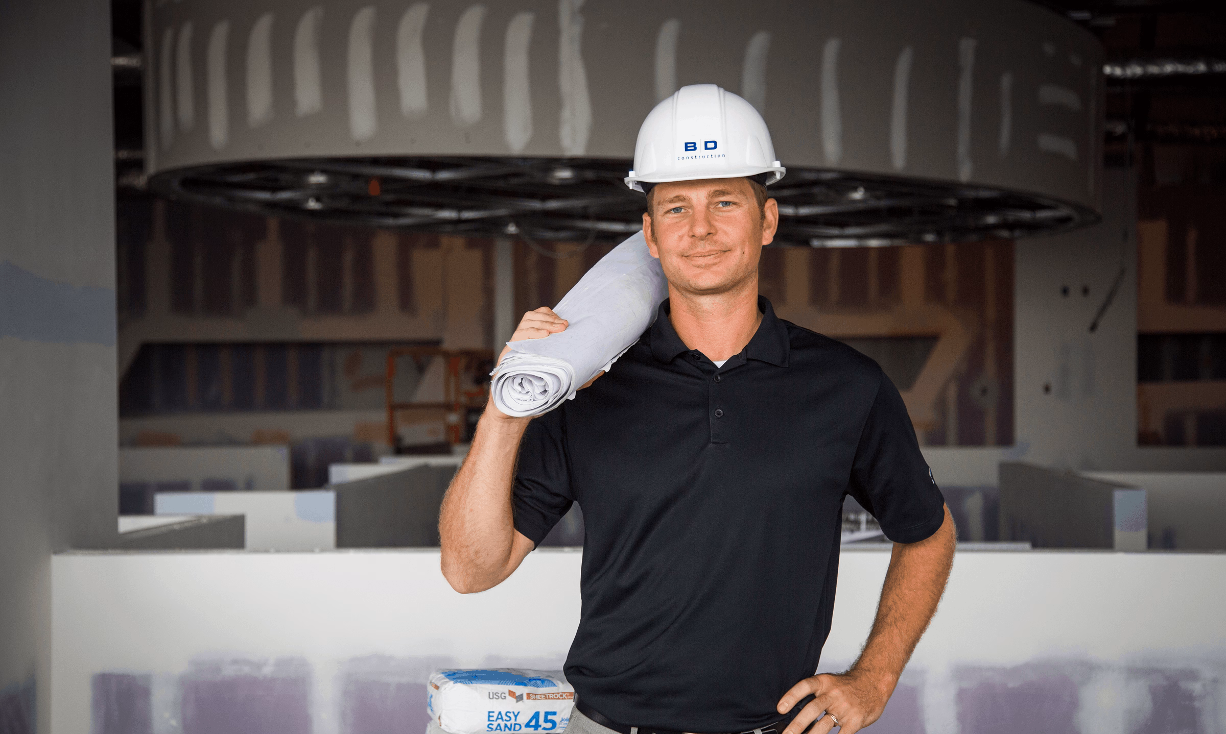 Man wearing a white construction helmet and black polo shirt standing inside a building under construction, holding rolled blueprints over his shoulder.