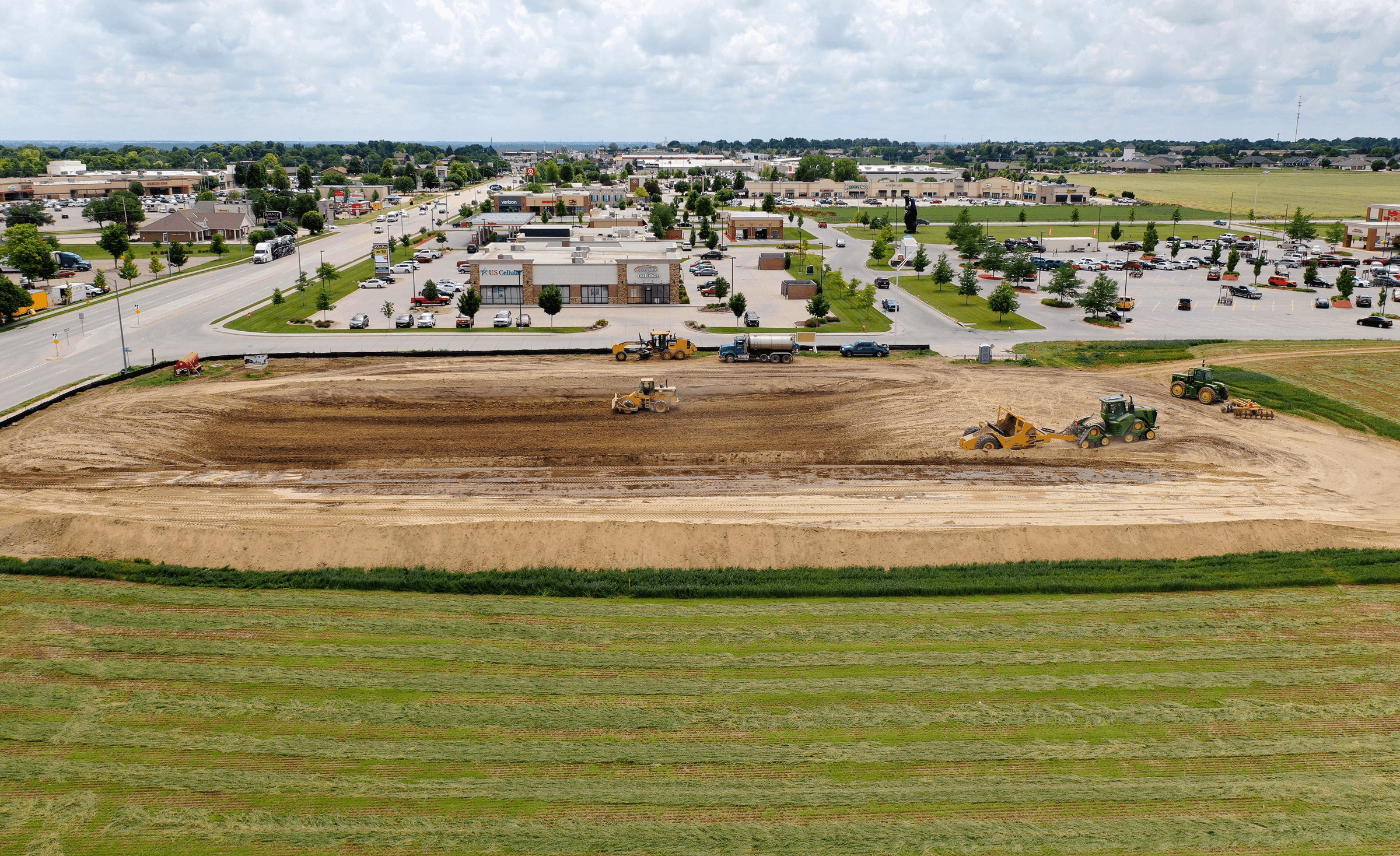 Aerial view of a construction site with heavy machinery leveling the ground, adjacent to a shopping plaza and parking lots under a partly cloudy sky.