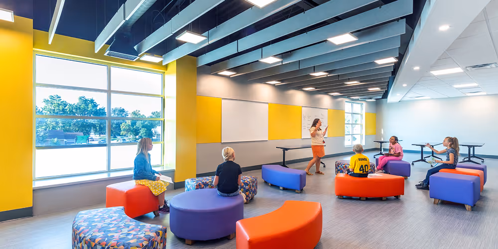 Modern classroom with colorful modular seating arranged in a semicircle, a teacher standing and speaking, and five students seated listening attentively.