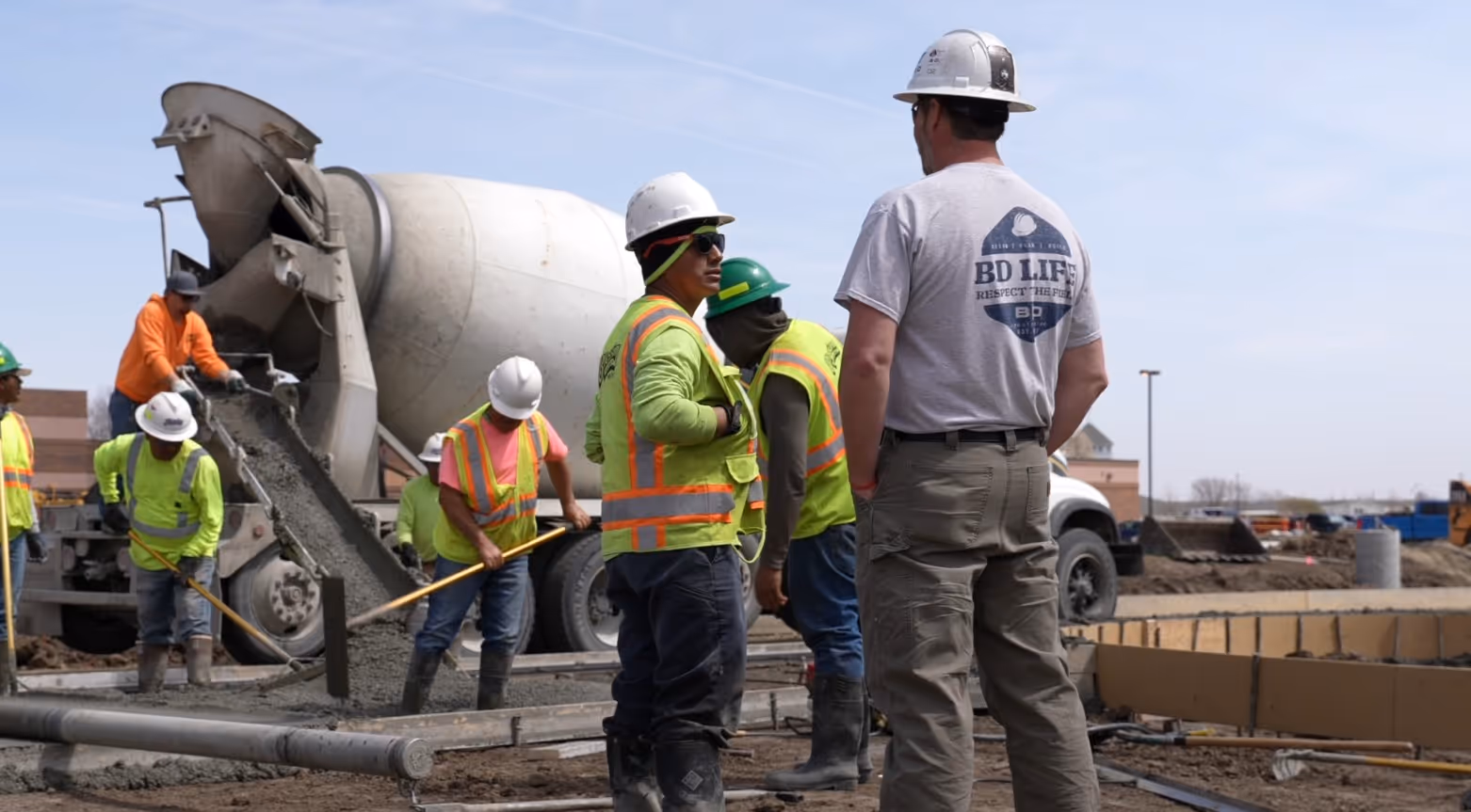 Construction workers in safety gear pour and spread concrete from a cement mixer on a building site.