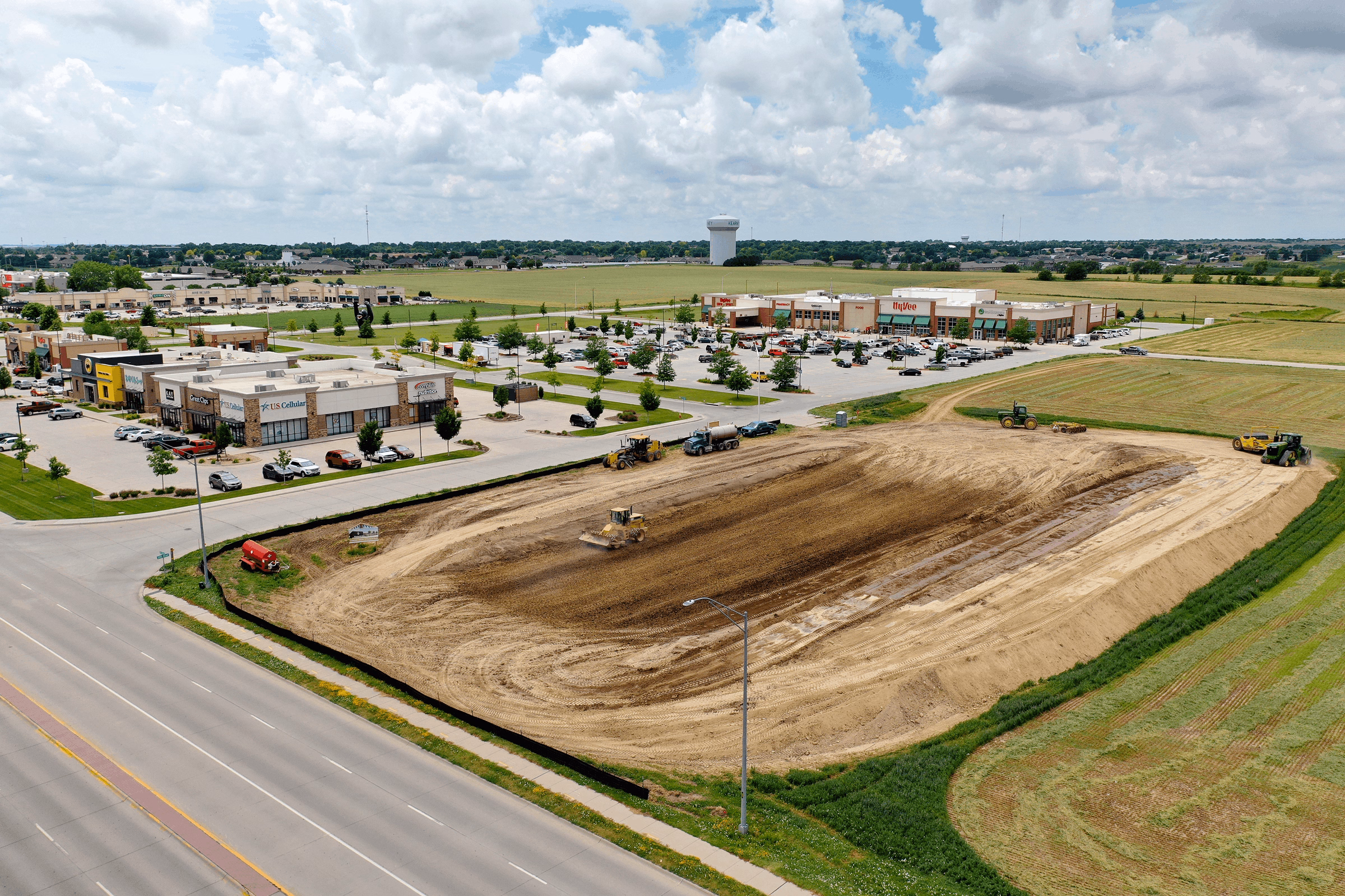 Aerial view of a construction site next to a shopping center with parked cars and surrounding green fields under a cloudy sky.