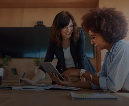 Two women smiling and discussing work with a tablet and notebooks at a desk.
