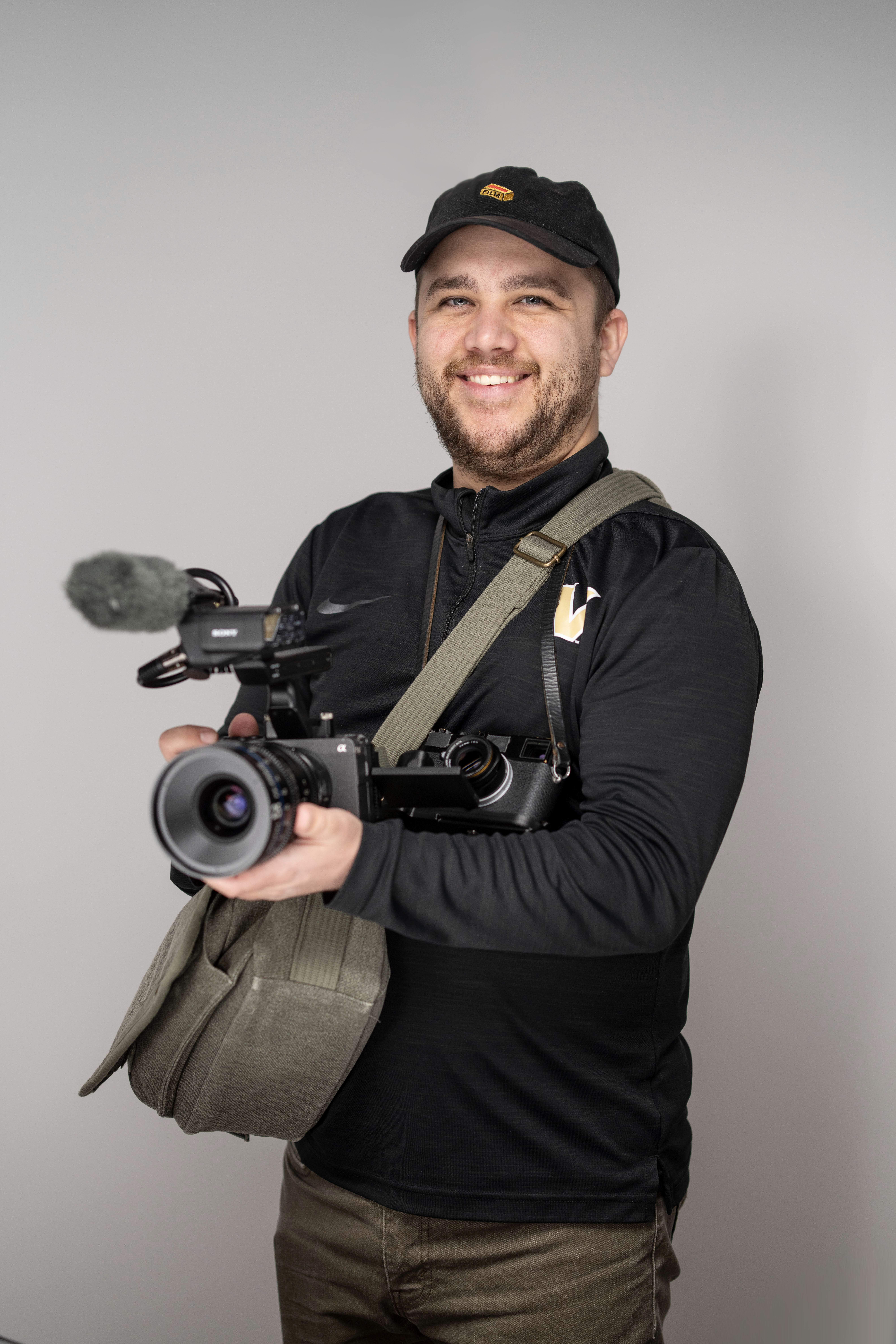 Alex smiling wearing a black cap holds a video camera with a microphone and carries a green bag.