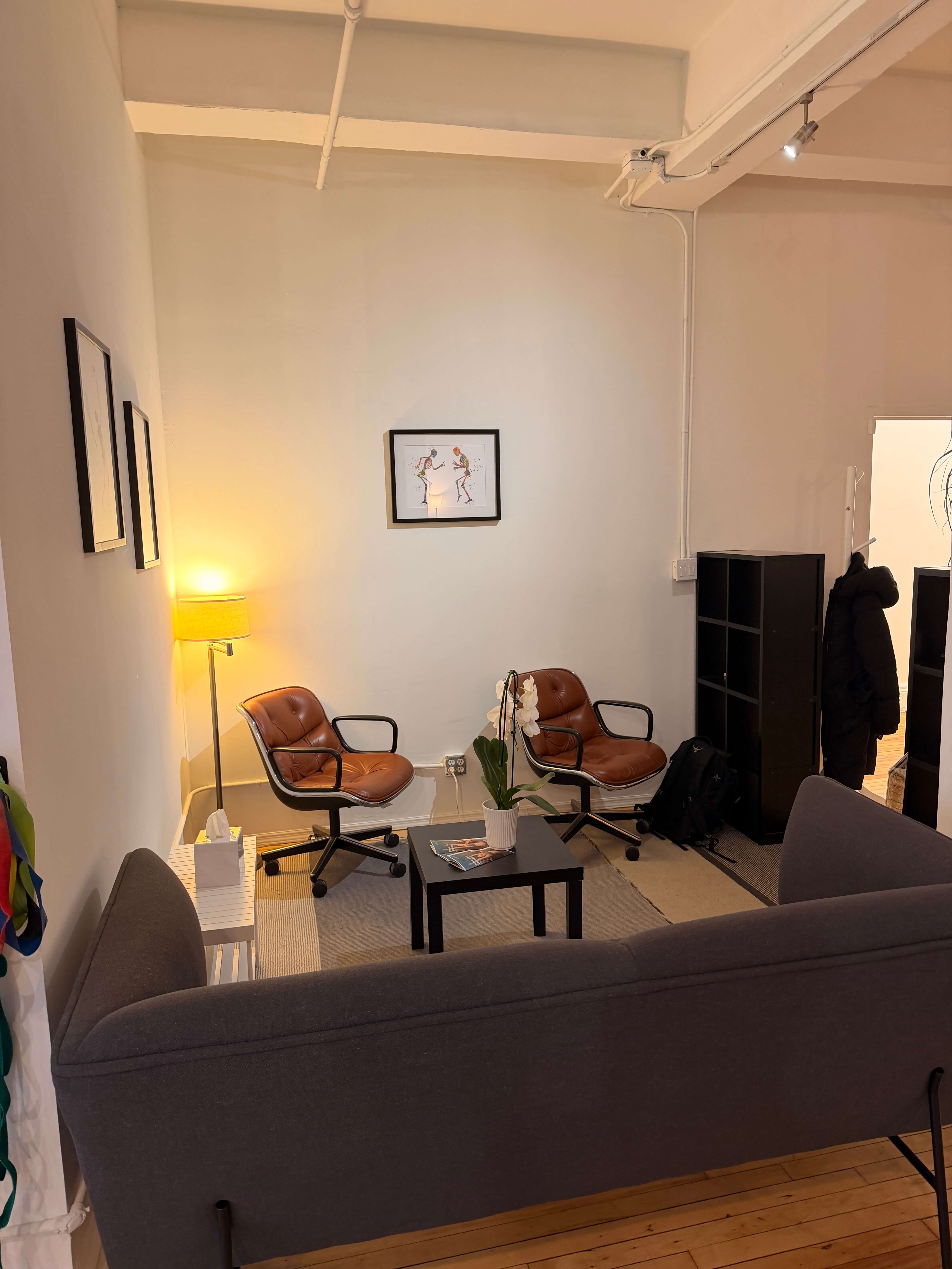 Cozy seating area with two brown leather chairs, a black coffee table with a white orchid, a gray couch, and a lit floor lamp against a white wall.