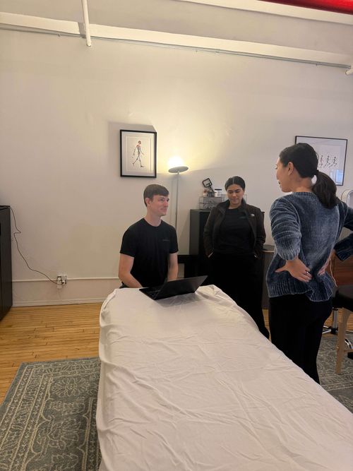 Three people in a room with a massage table; one man sits behind a laptop on the table, two women stand nearby talking.