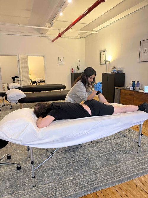 Therapist wearing blue gloves stretching the foot of a person lying face down on a massage table in a treatment room.