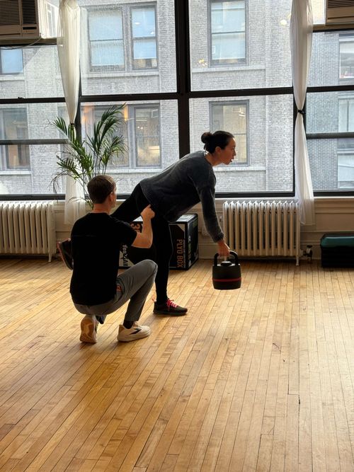 A woman holding a kettlebell in one hand while balancing on one leg, assisted by a man kneeling beside her in a spacious room with large windows and wooden floors.