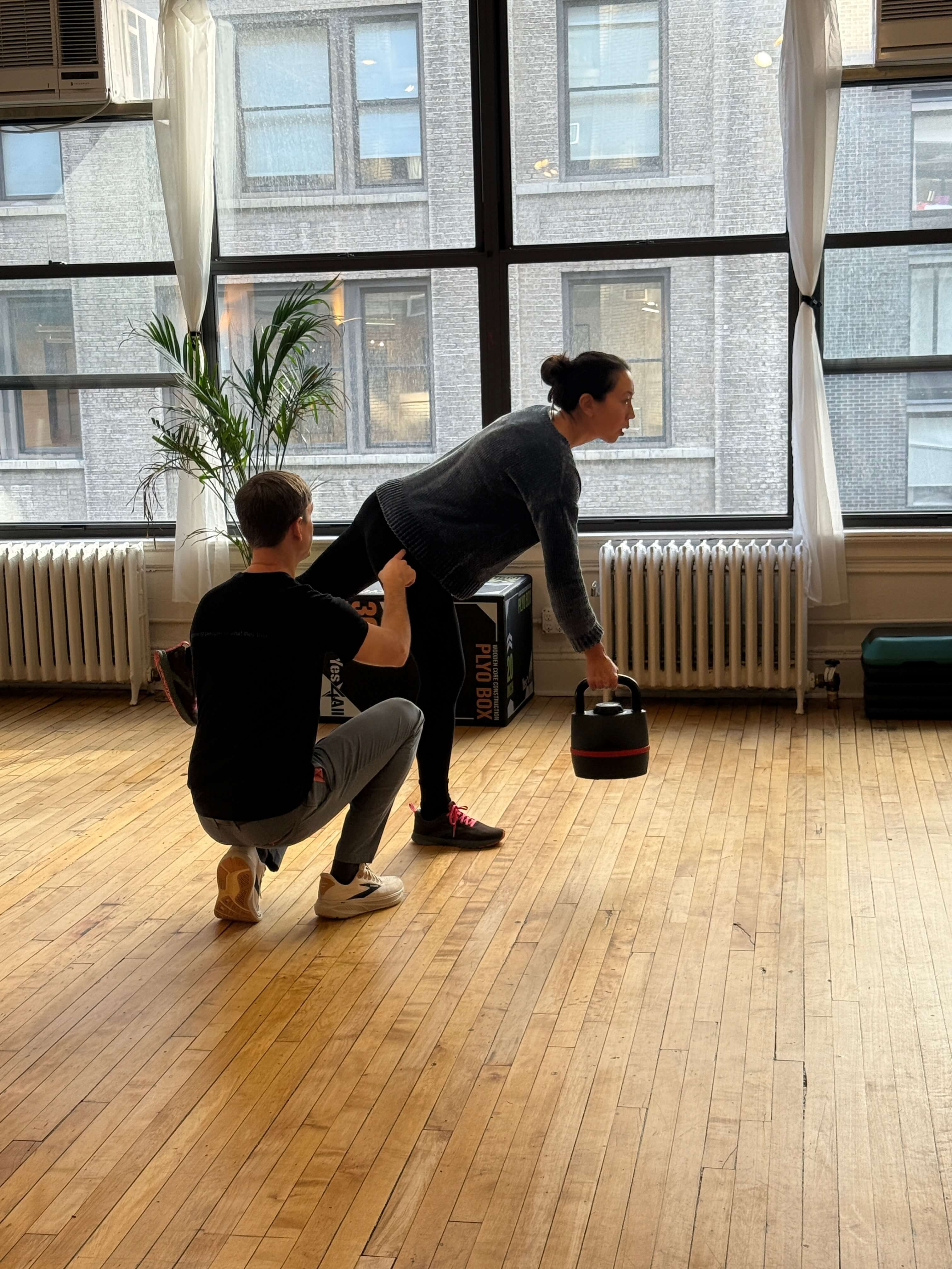A woman holding a kettlebell in one hand while balancing on one leg, assisted by a man kneeling beside her in a spacious room with large windows and wooden floors.