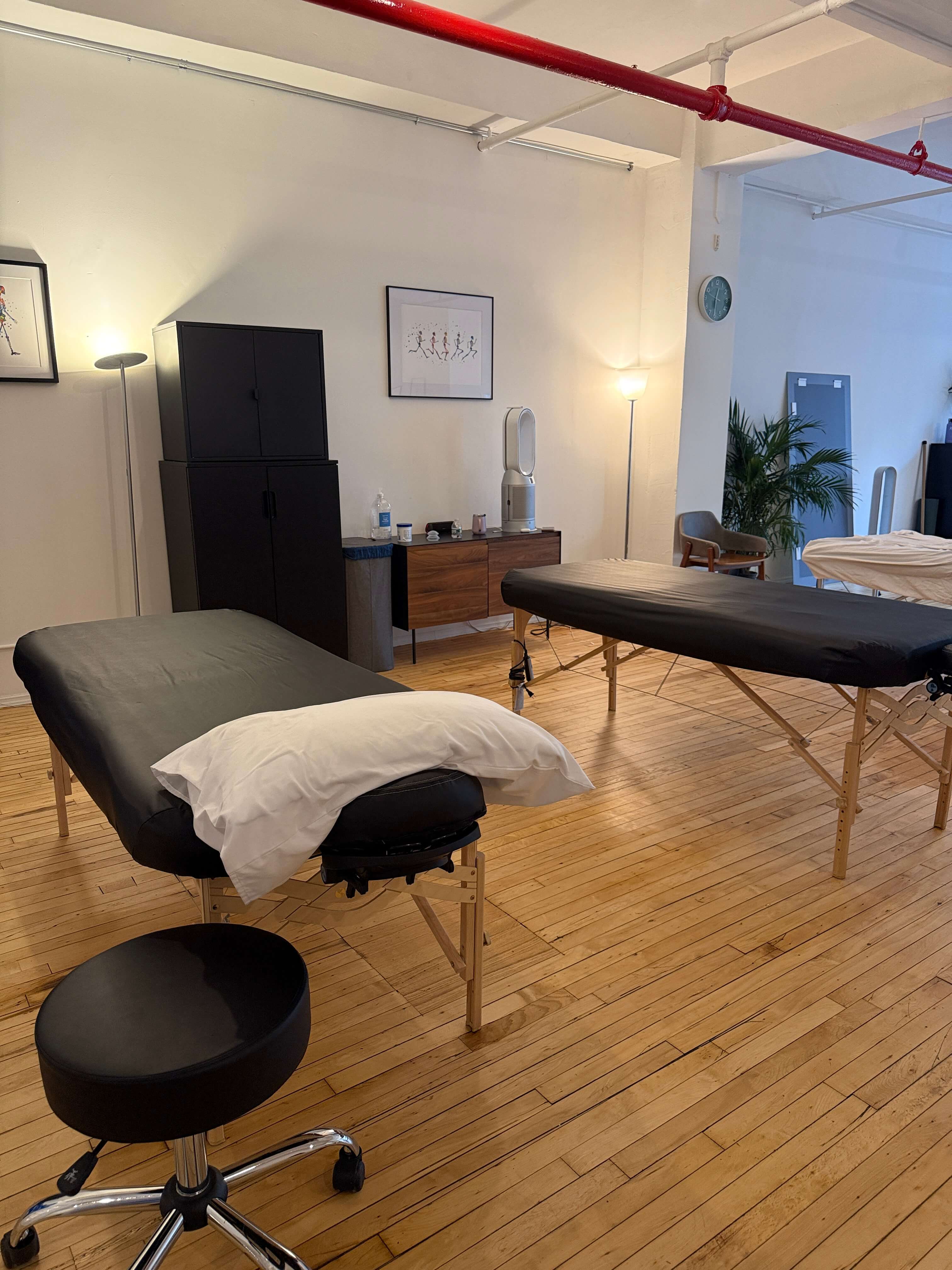 Massage therapy room with two black-covered massage tables, a white pillow, a black stool, wooden floor, and minimalistic decor including lamps and framed artwork.