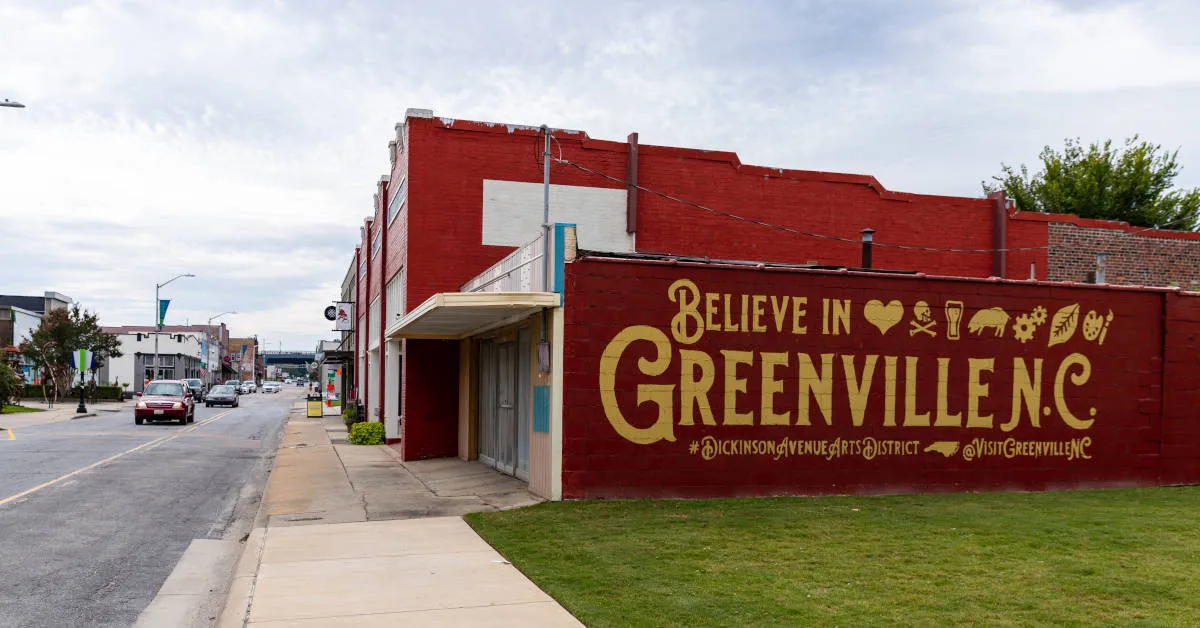 Street view of a red brick building with a large yellow mural reading 'Believe in Greenville N.C.' accompanied by icons and hashtags.